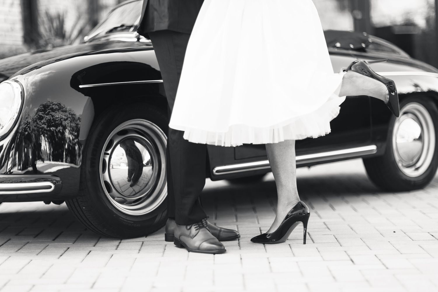Black and white photo of bride and groom standing beside a classic vintage Porsche convertible.