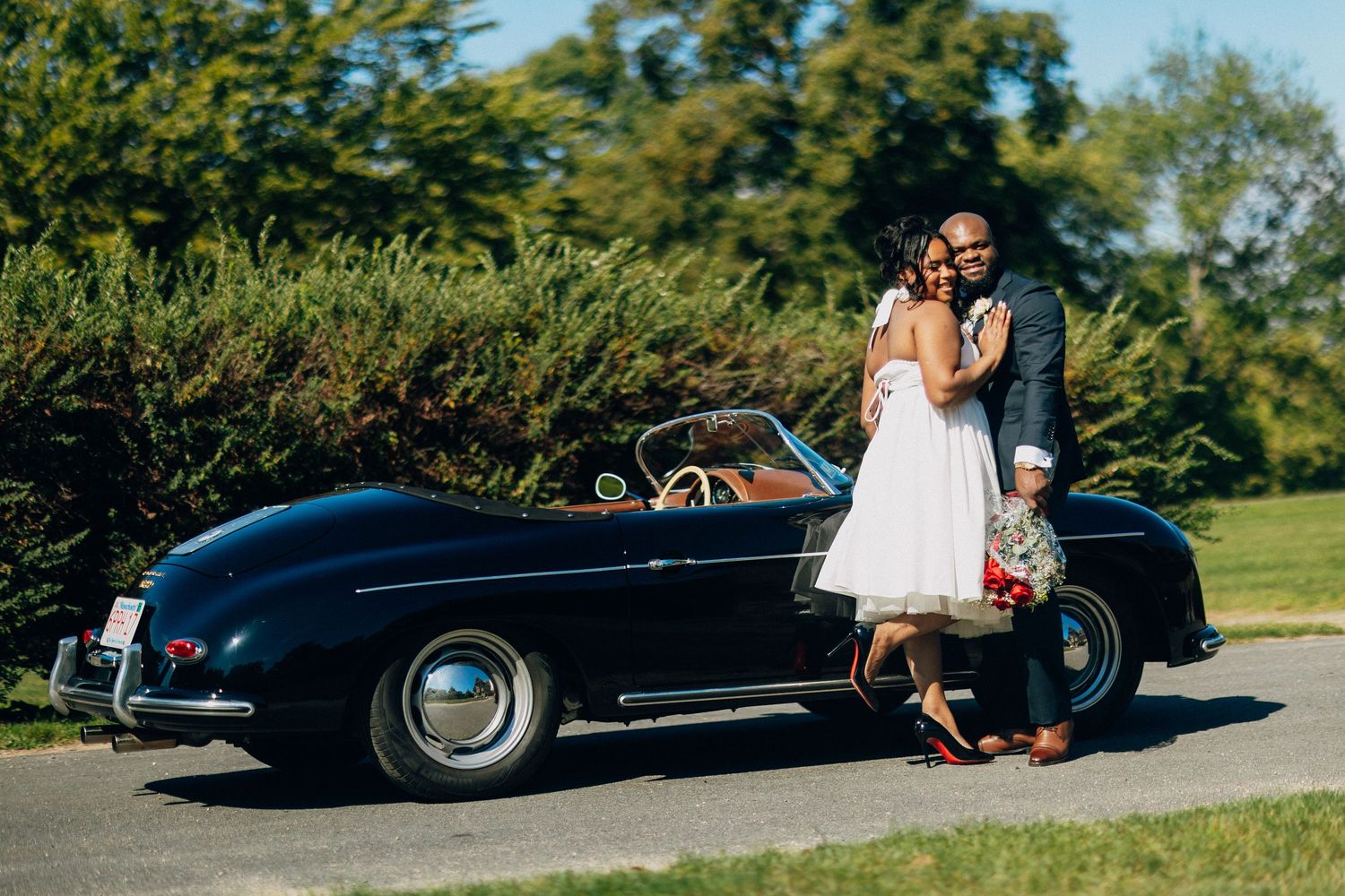 A smiling couple embraces beside a black vintage convertible on a sunny tree-lined road.