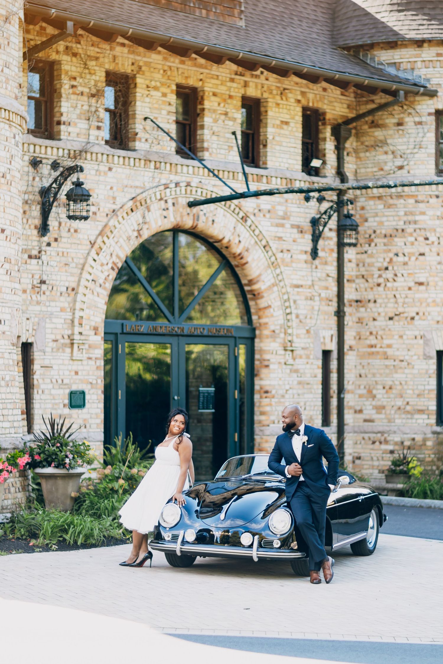 Elegant couple in wedding attire leaning against a classic black sports car outside a brick building.