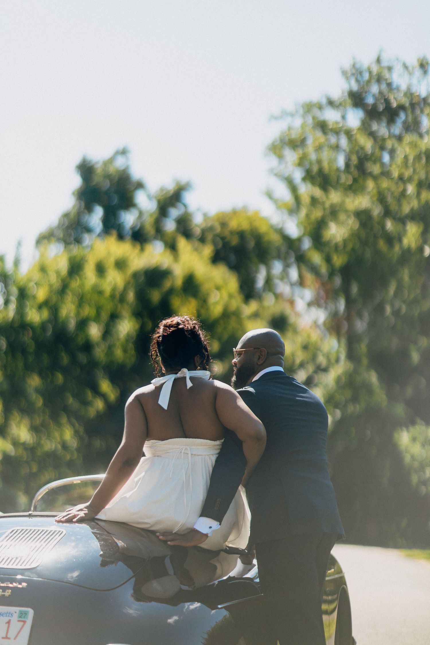 Bride and groom lean against a vintage car hood, viewed from behind, surrounded by lush greenery.
