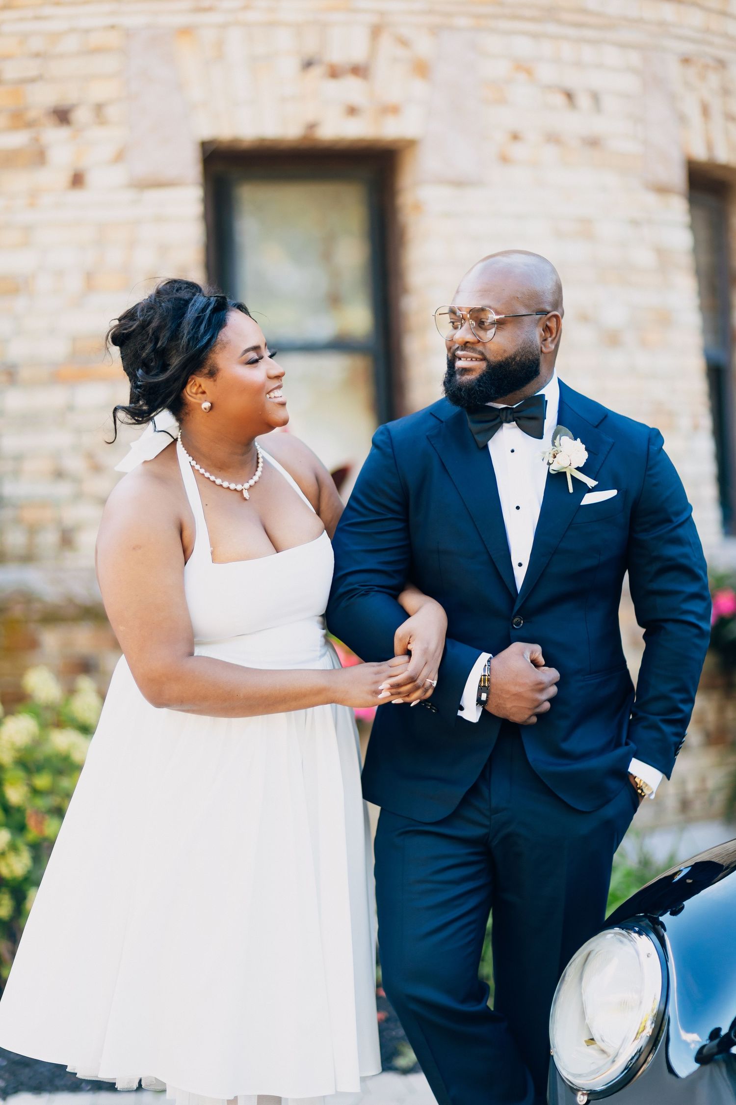 Bride in white halter dress and groom in navy tuxedo walking together near a classic car.
