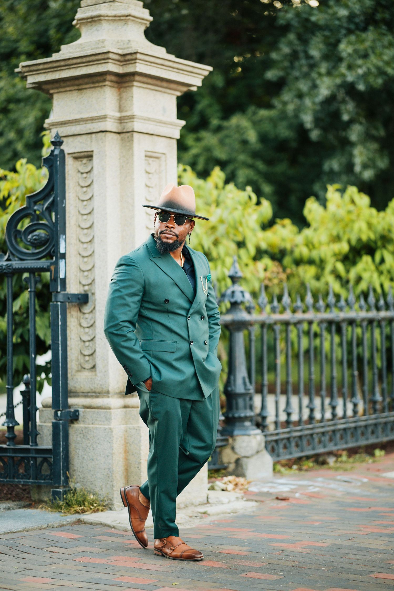 Stylish man in green double-breasted suit and hat leaning against stone pillar.