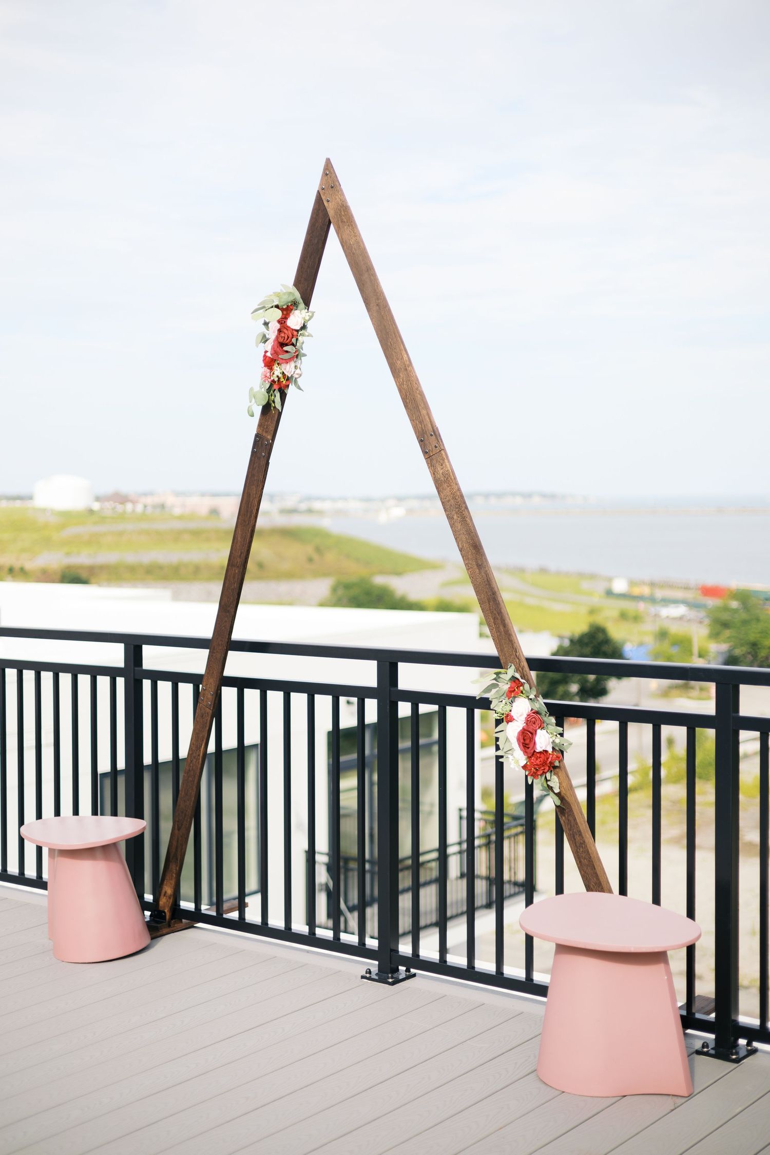 Triangular wooden wedding arch with floral accents on rooftop deck, pink stools, overlooking waterfront.