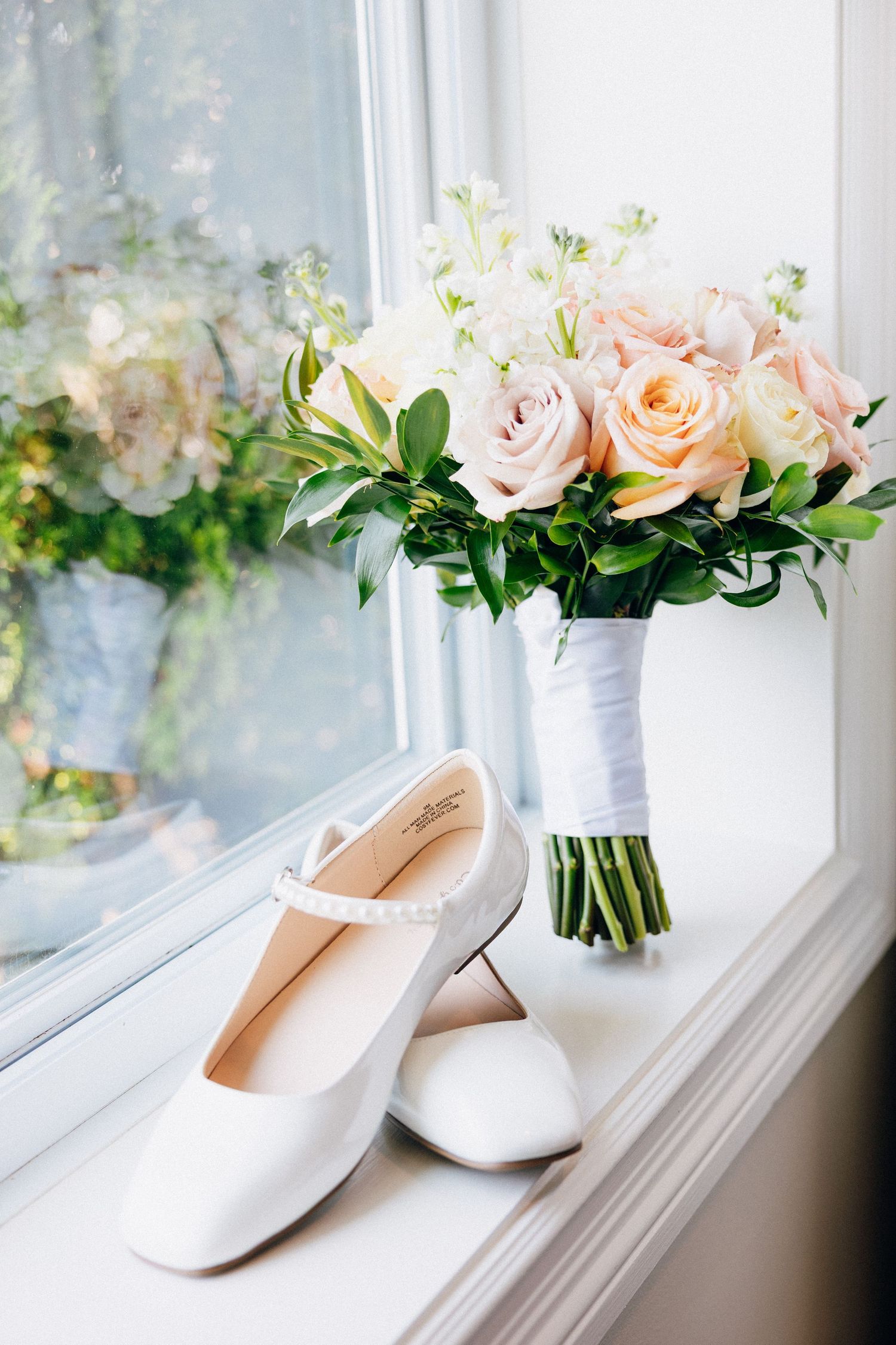 White bridal heels and a blush pink rose bouquet with greenery displayed on a sunlit window sill.