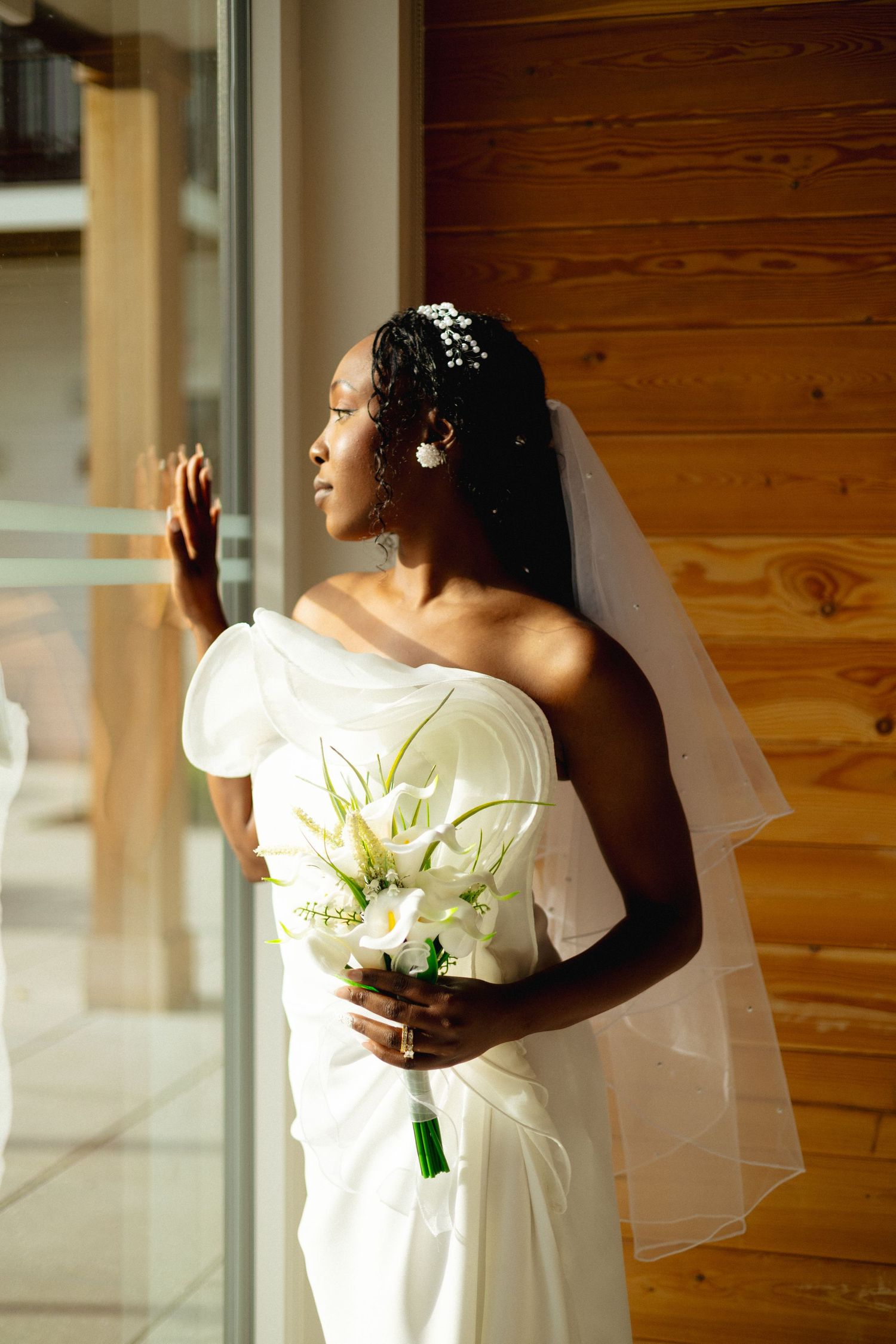 Beautiful Black bride in white strapless gown and veil holds white calla lily bouquet by window.