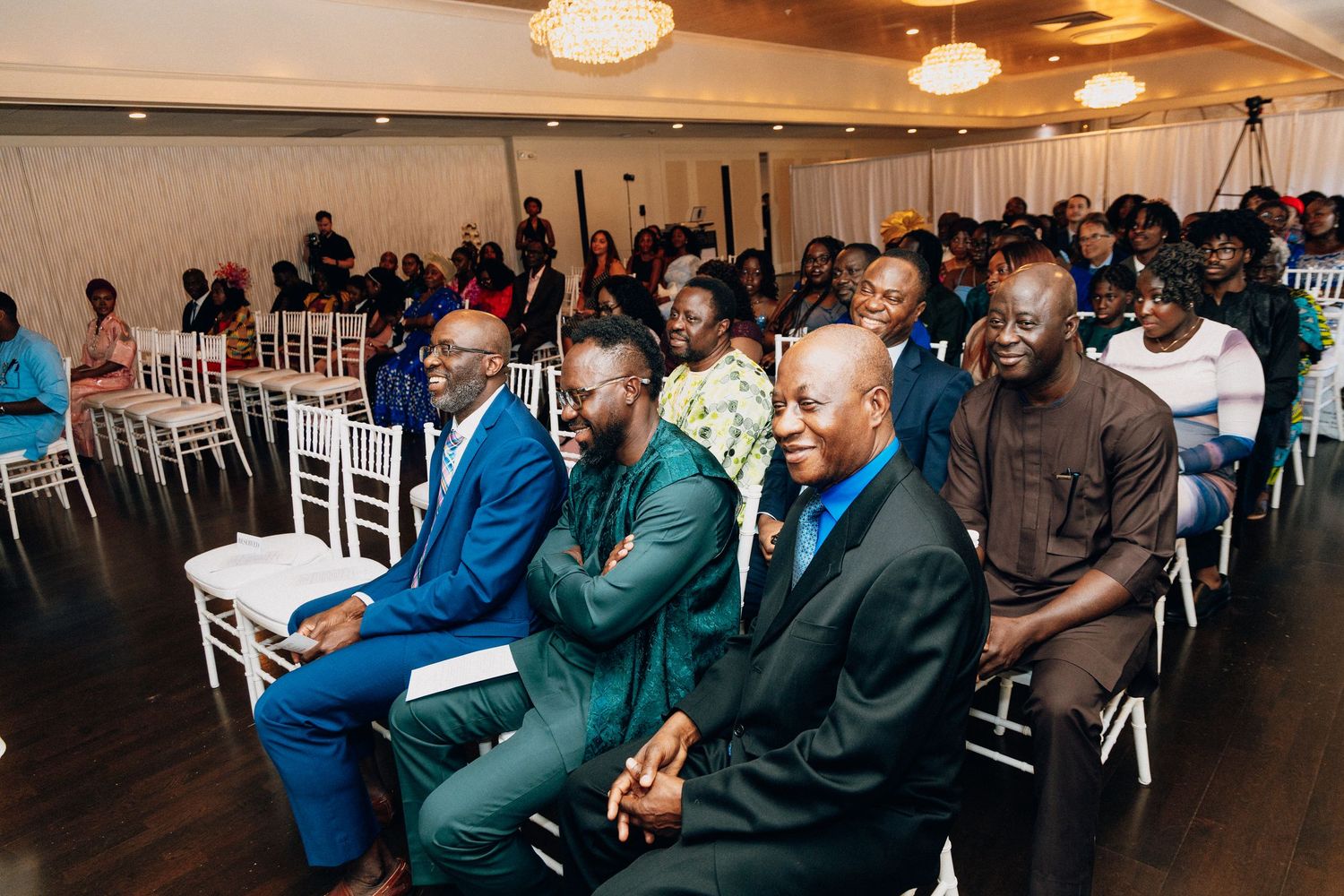 Diverse group of elegantly dressed guests seated in rows at a formal indoor event or ceremony.