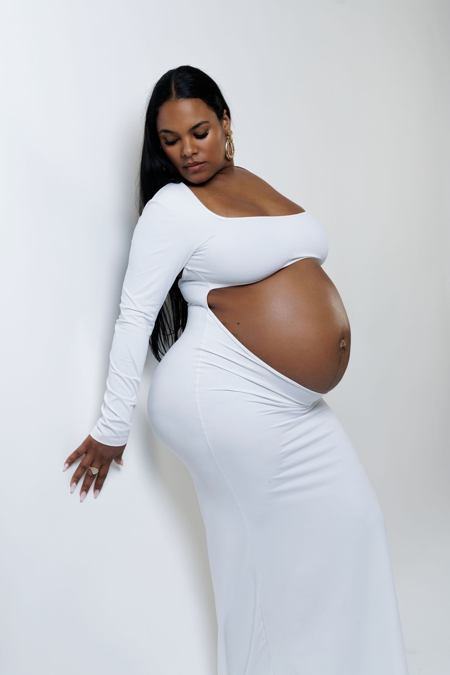 Pregnant woman in white cutout maxi dress posing against white wall, showcasing baby bump in elegant maternity photo.