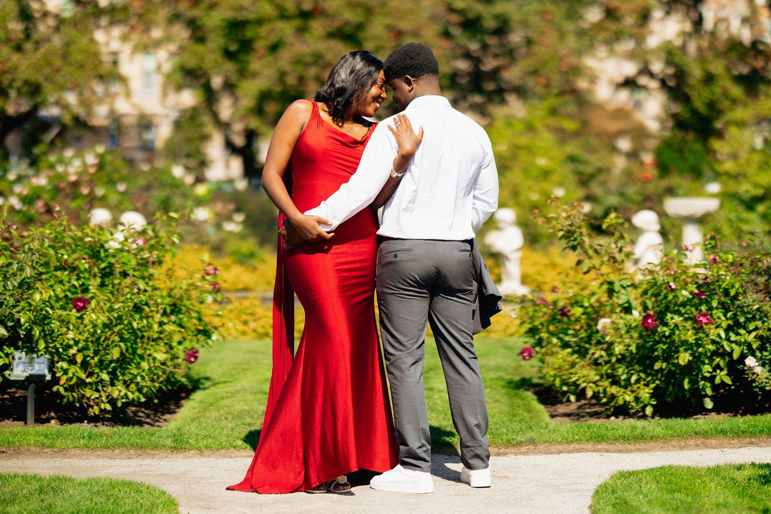 Couple in red dress and white shirt embracing in a lush garden with colorful flowers.