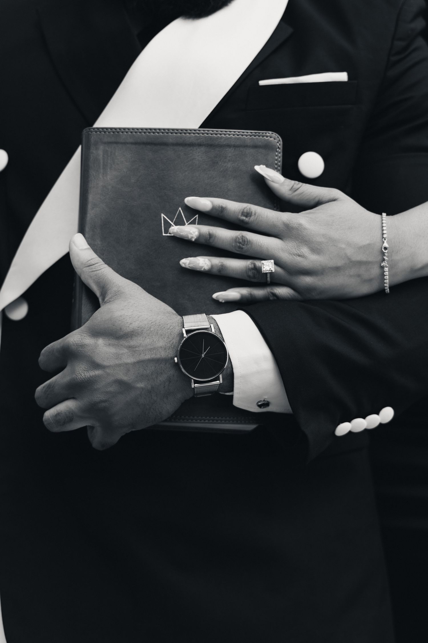Black and white photo of hands holding a leather notebook, man wearing suit and watch, woman wearing bracelet and ring.