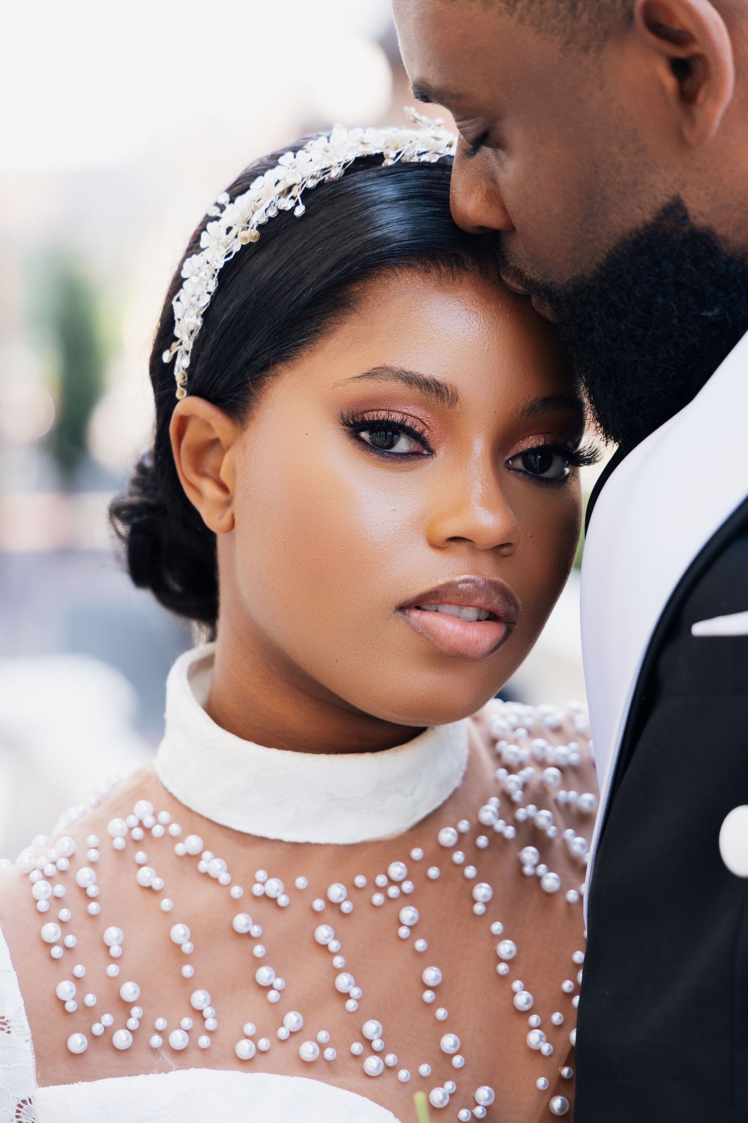 A Black bride in a pearl-embellished white gown and crystal headband gazes forward as her groom kisses her forehead.