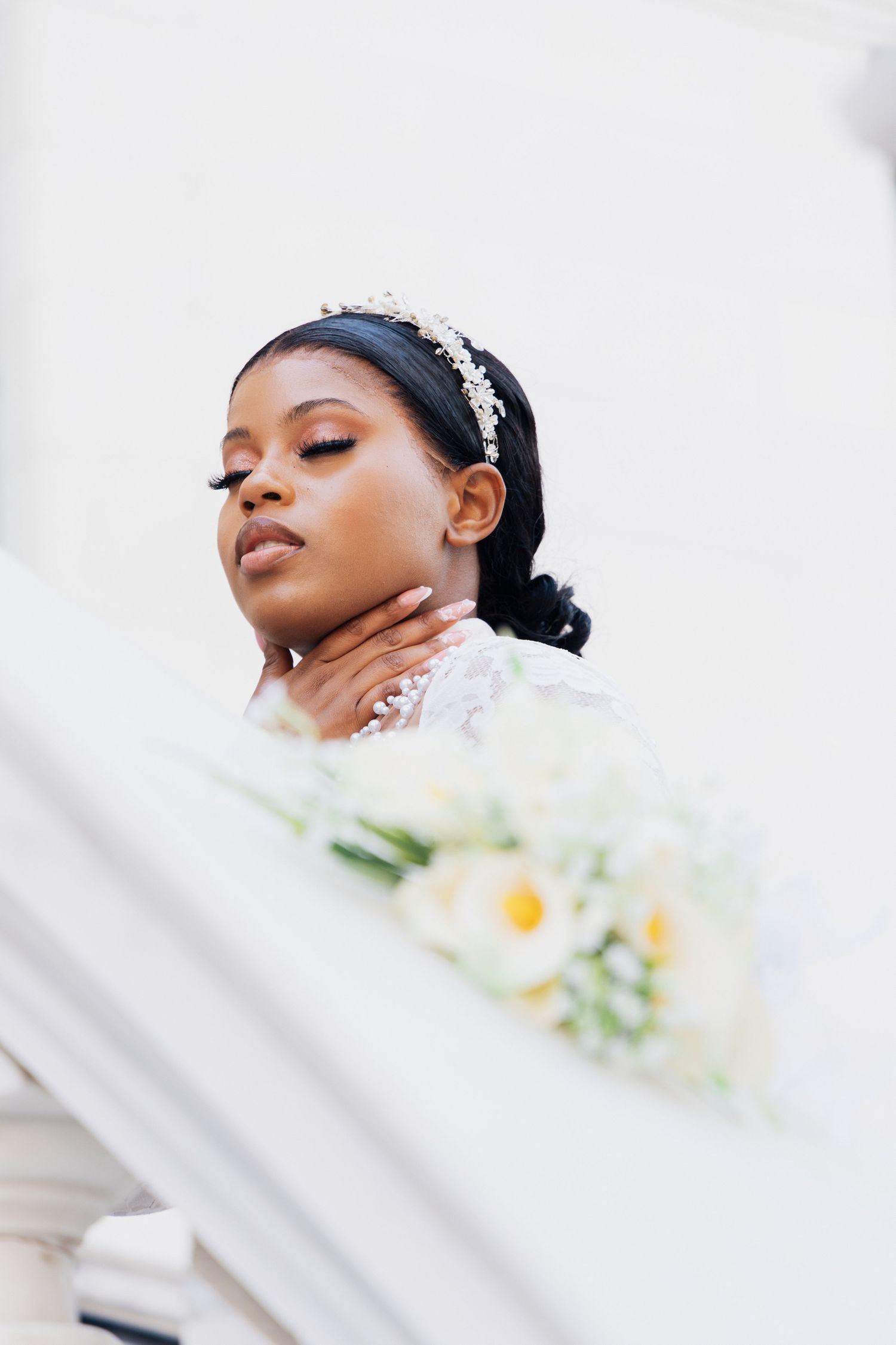 A Black bride in a white lace gown and crystal tiara holds a white floral bouquet while posing gracefully outdoors.