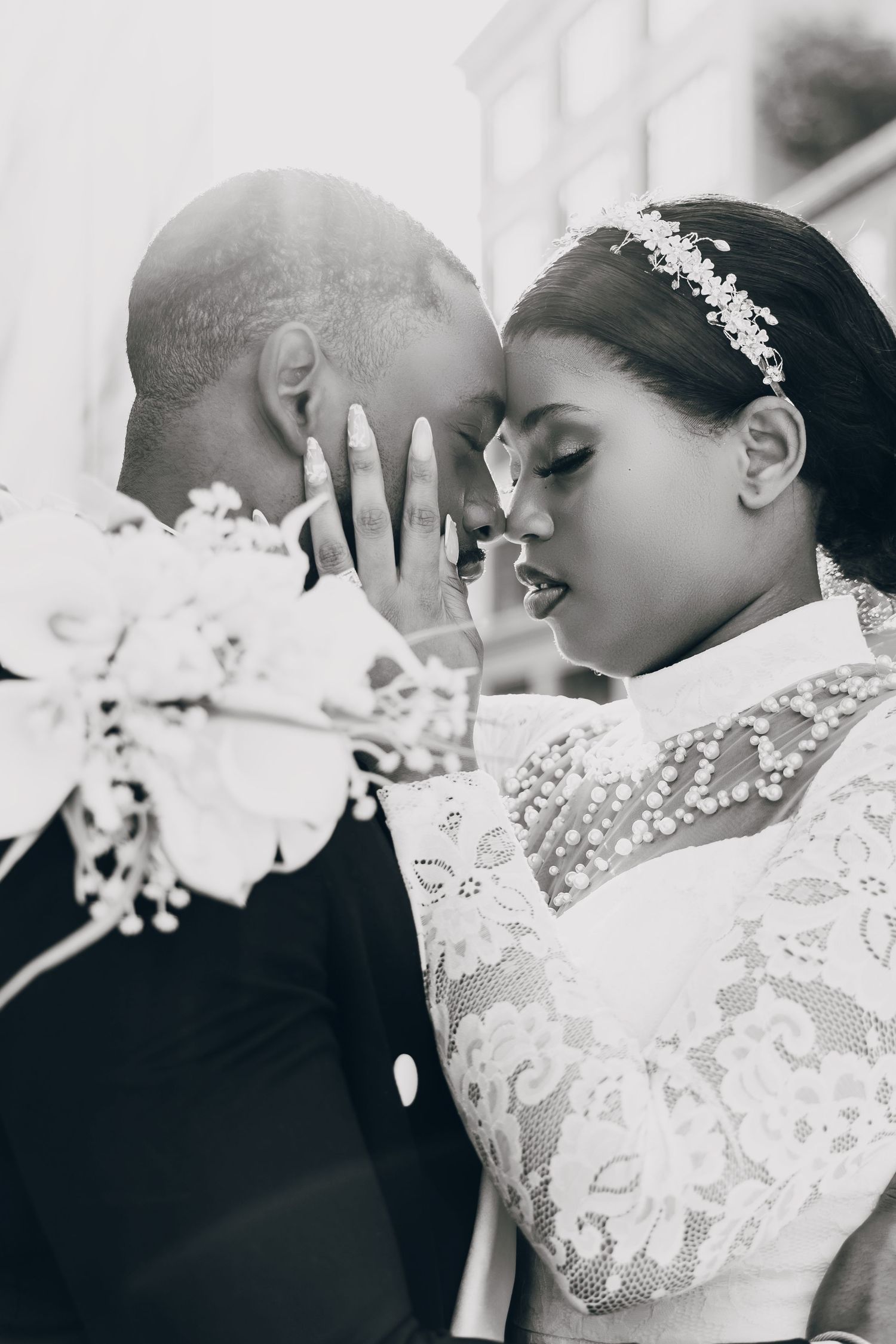 Black and white photo of a bride and groom touching foreheads, bride in lace gown with floral headpiece holding bouquet.