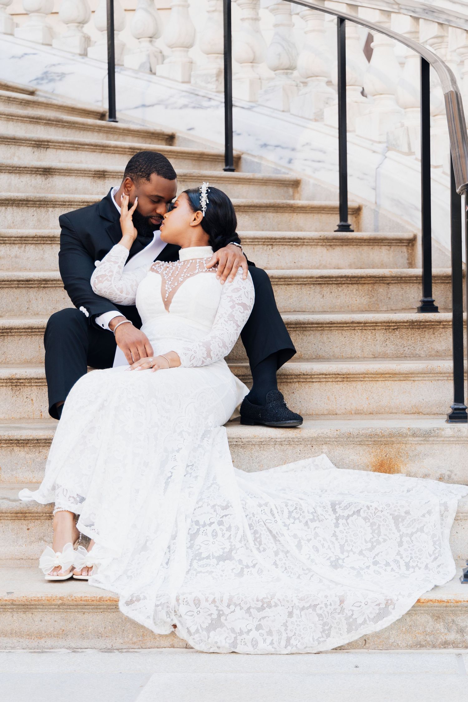 Bride and groom share a romantic kiss on marble steps, bride wearing elegant long-sleeve lace wedding gown.
