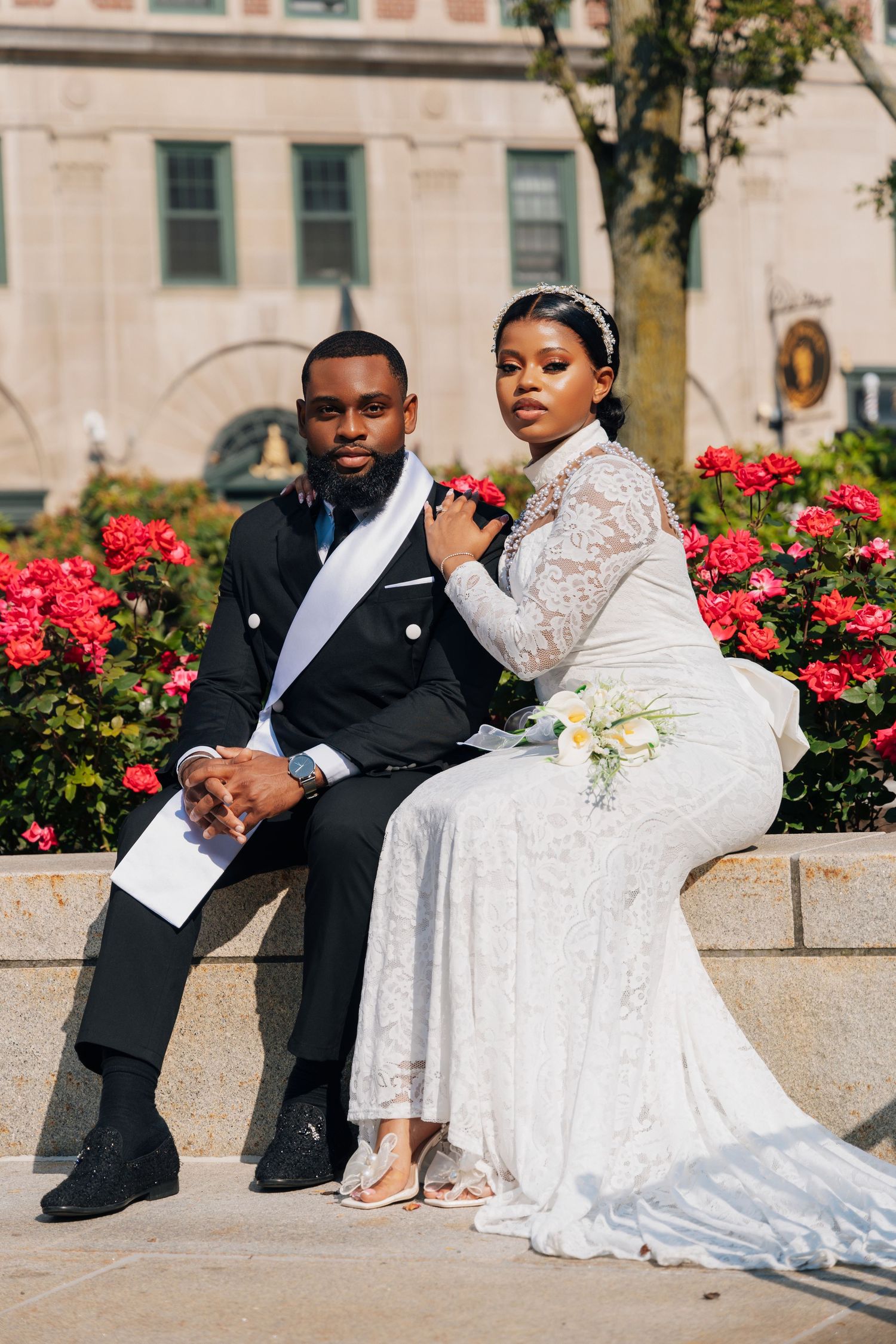 A Black couple in wedding attire sits outdoors by red flowers, the bride in white lace gown and groom in black tuxedo.