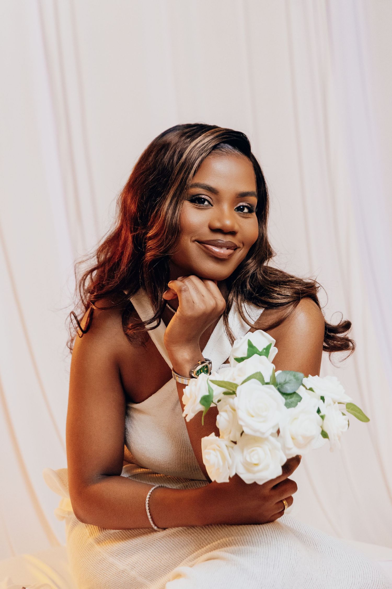 Beautiful Black woman smiling softly, holding white roses bouquet in a chic white sleeveless dress.