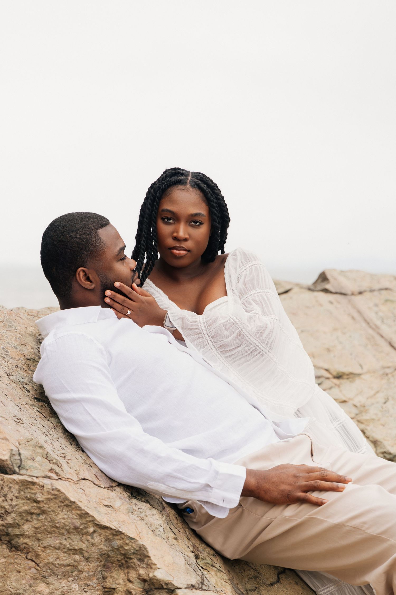 A tender moment as a woman with braids gently touches a mans face while reclining on rocks together outdoors.