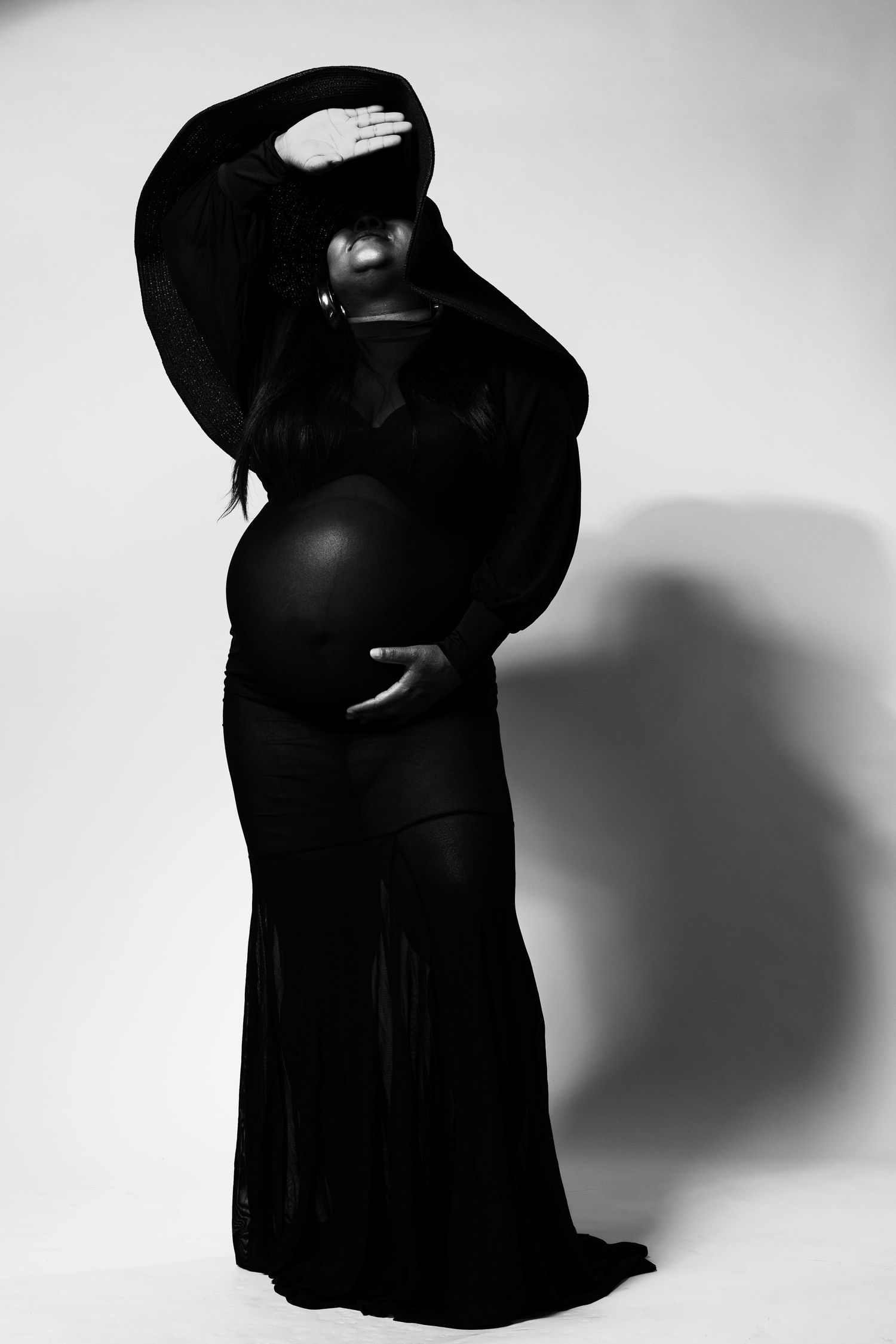Black and white maternity photo of pregnant woman in elegant black gown cradling baby bump in studio.