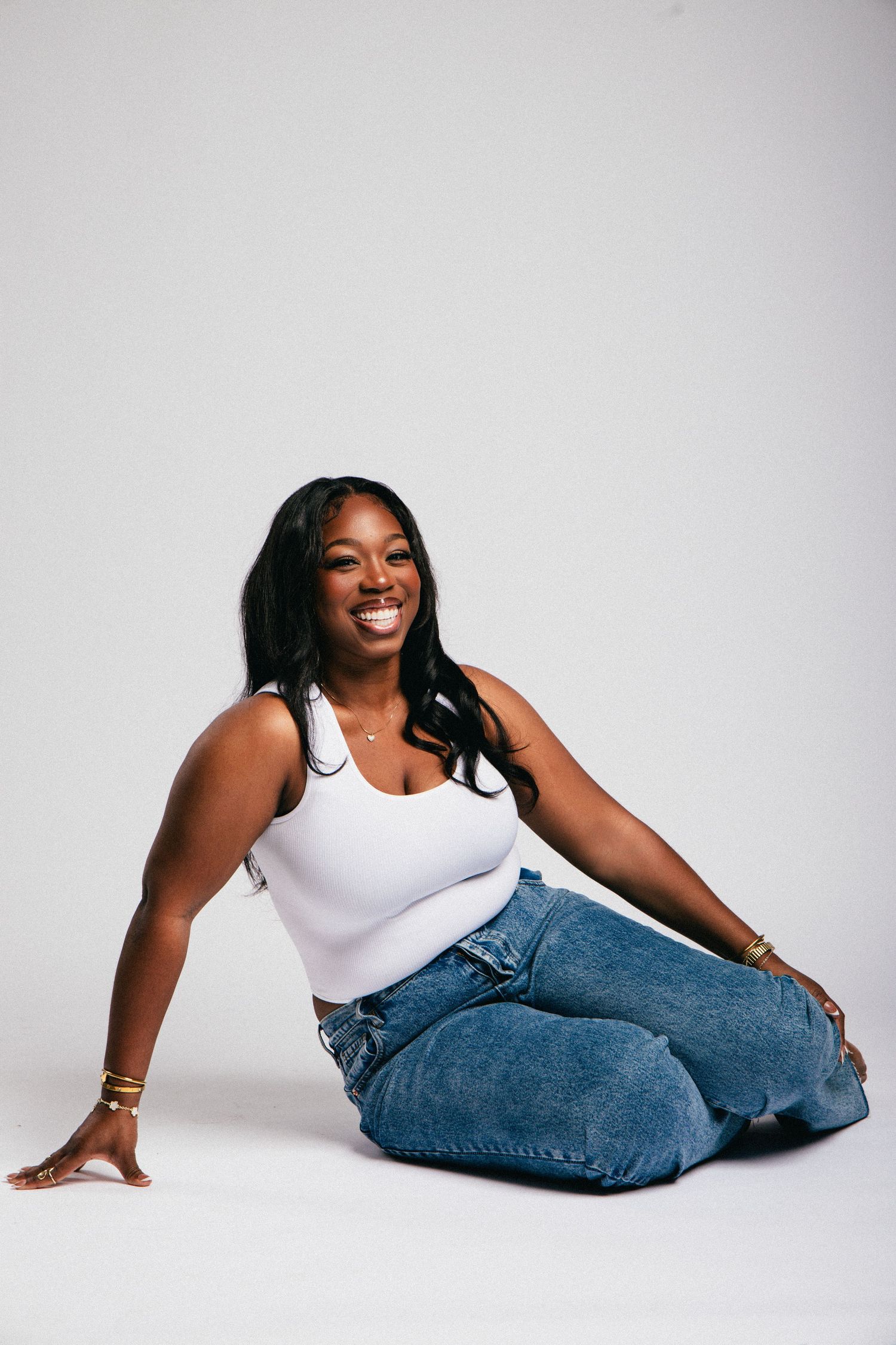 Smiling Black woman in white tank top and blue jeans sitting on floor in studio setting.