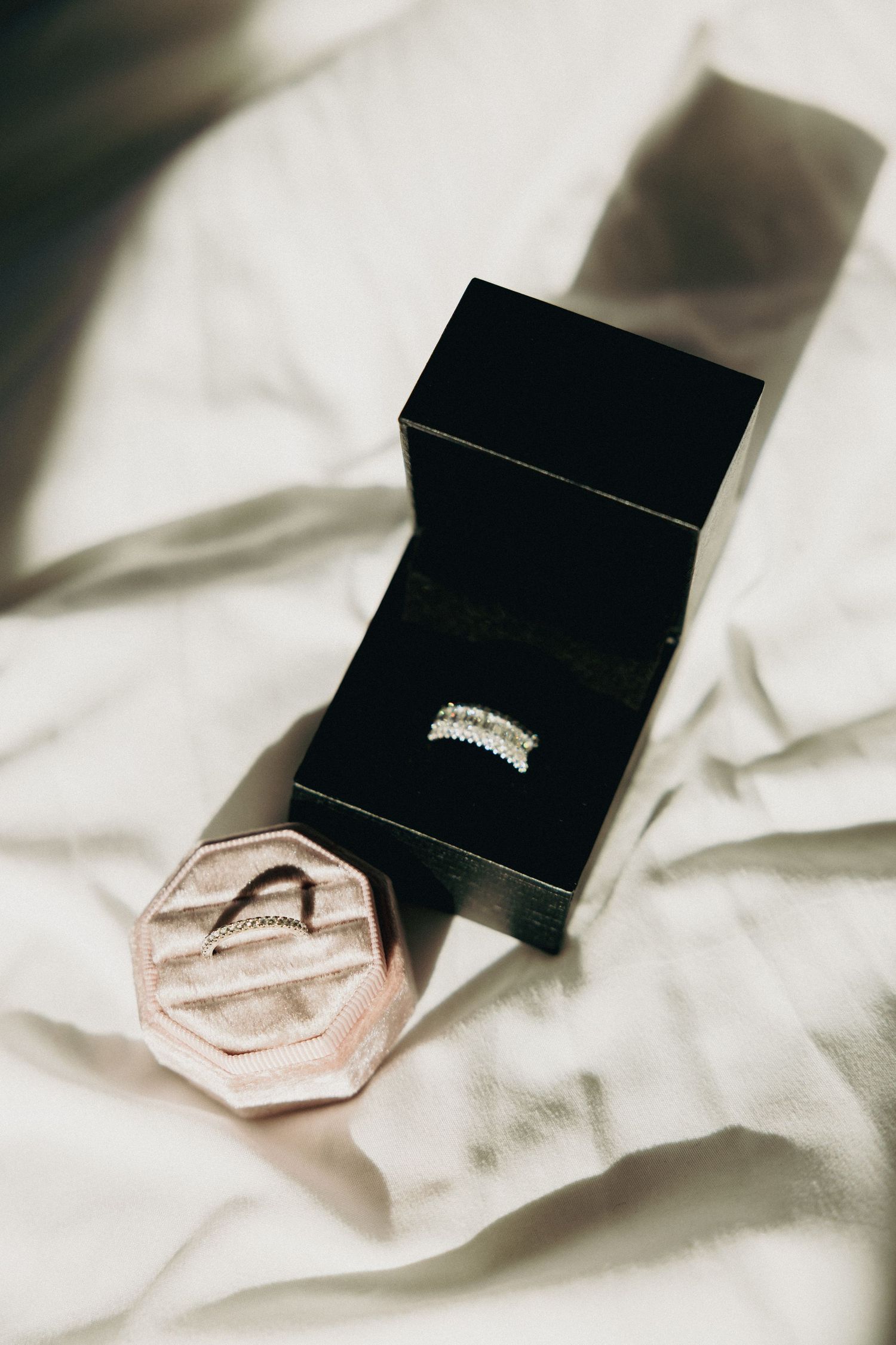 Diamond wedding band displayed in black ring box beside pink velvet ring box on white fabric.