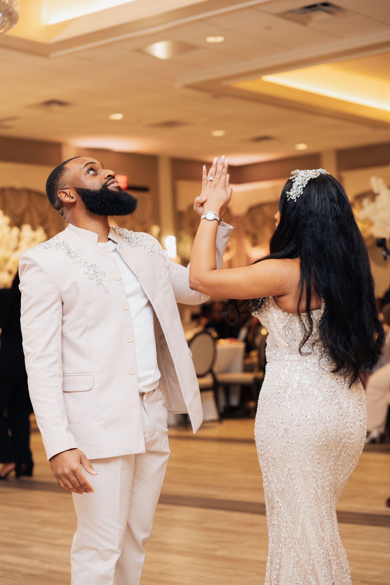 Bride and groom share joyful first dance in elegant ballroom, groom in white suit, bride in beaded gown.