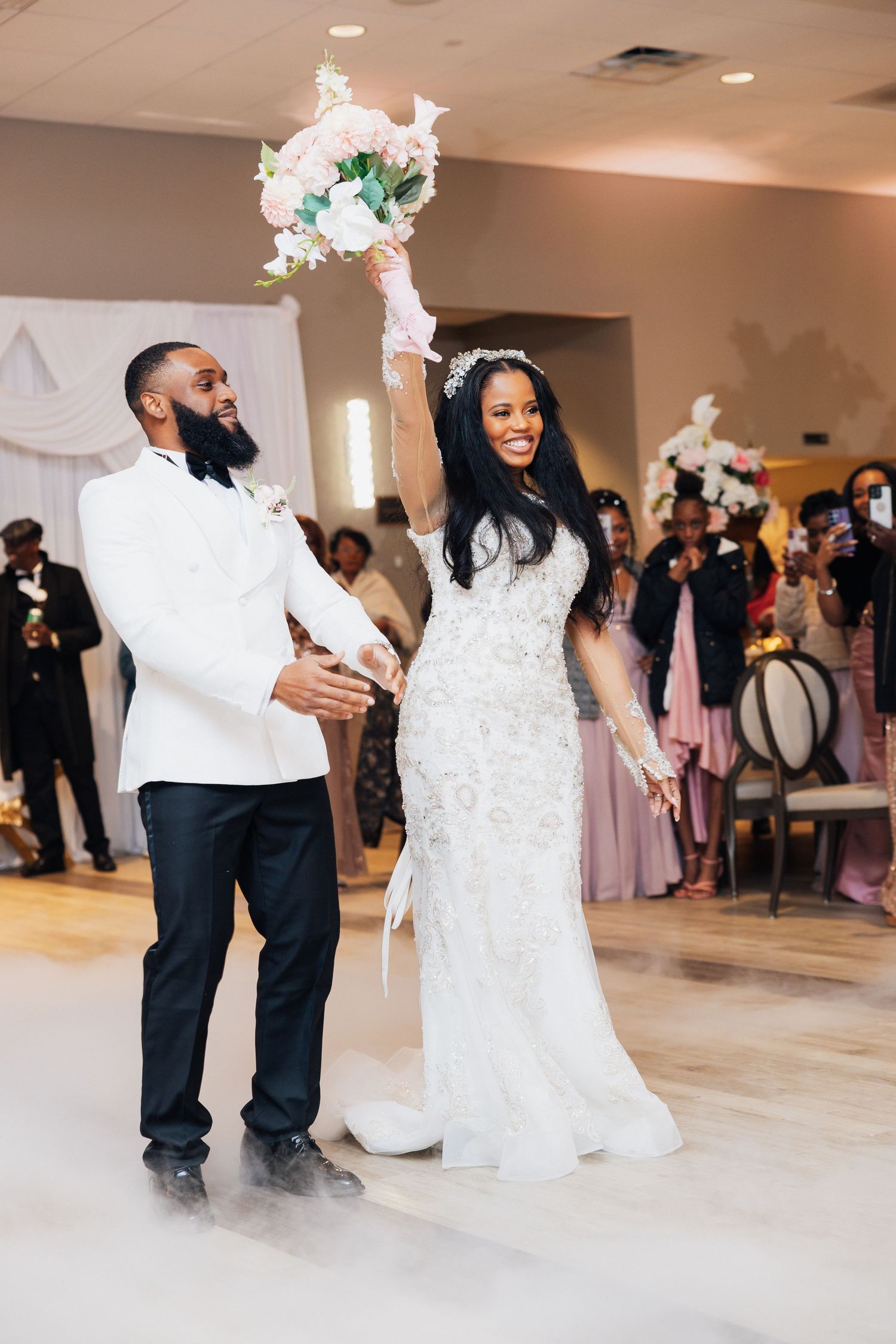 Joyful bride raises bouquet while groom holds her hand during wedding reception grand entrance with fog effect.