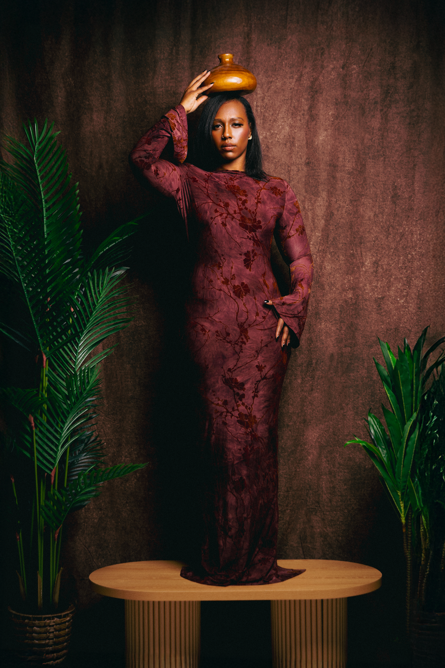 Woman in burgundy lace gown balancing gold vase on head, standing on table surrounded by tropical plants.