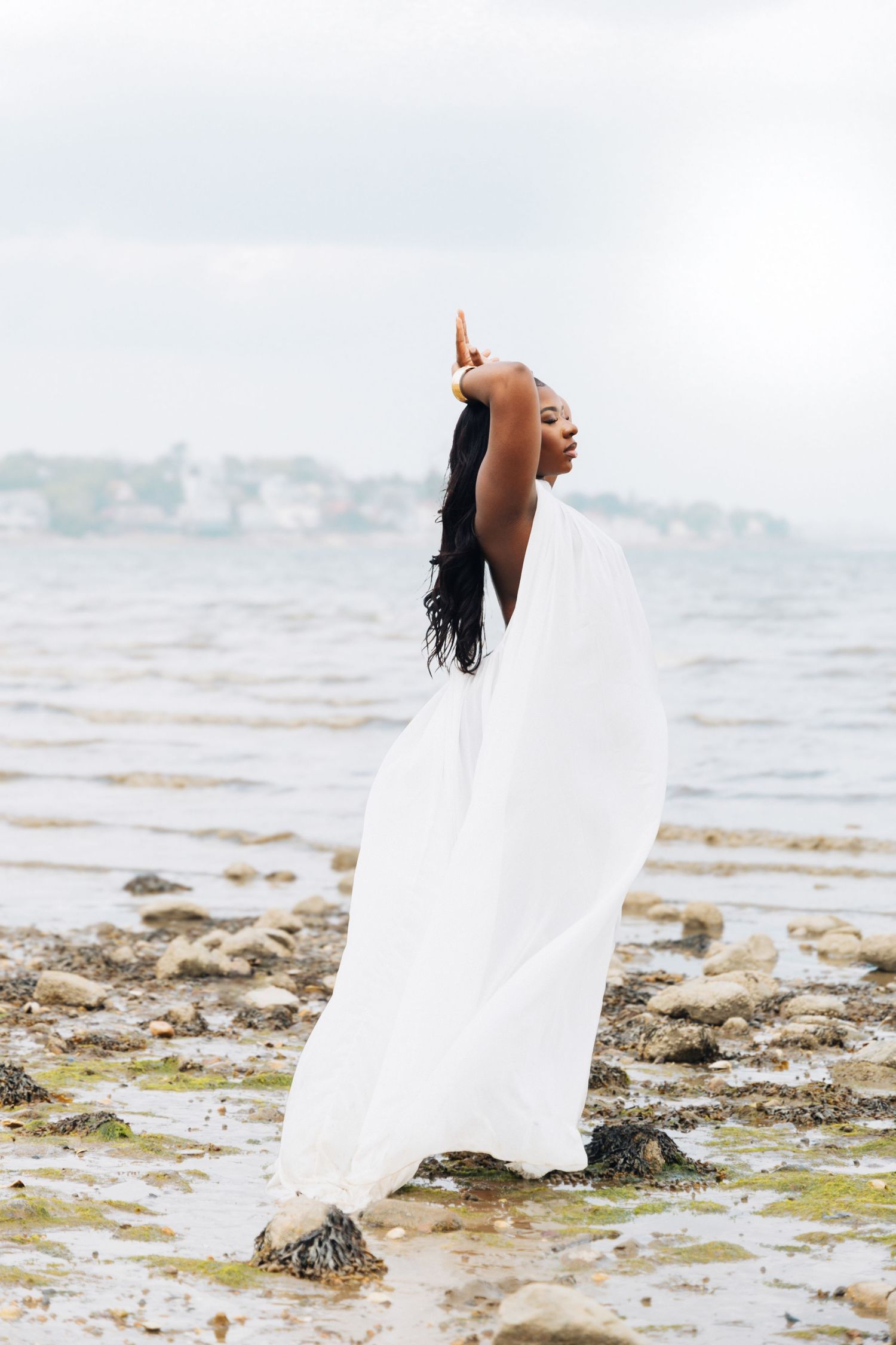 A Black woman in a flowing white fabric poses gracefully on rocky shoreline with ocean backdrop.