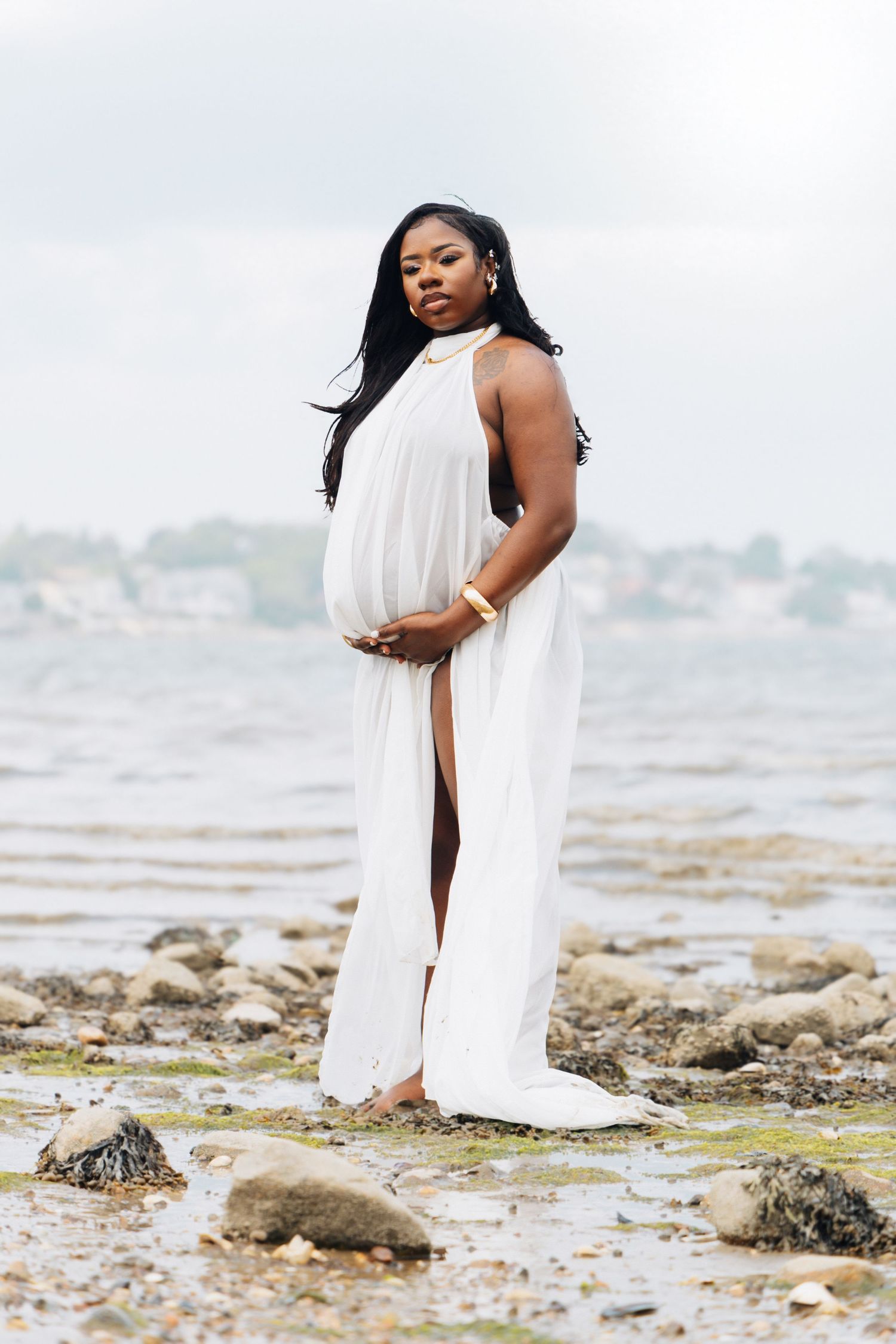 Pregnant Black woman in flowing white halter dress standing on rocky shoreline, cradling baby bump with serene coastal backdrop.
