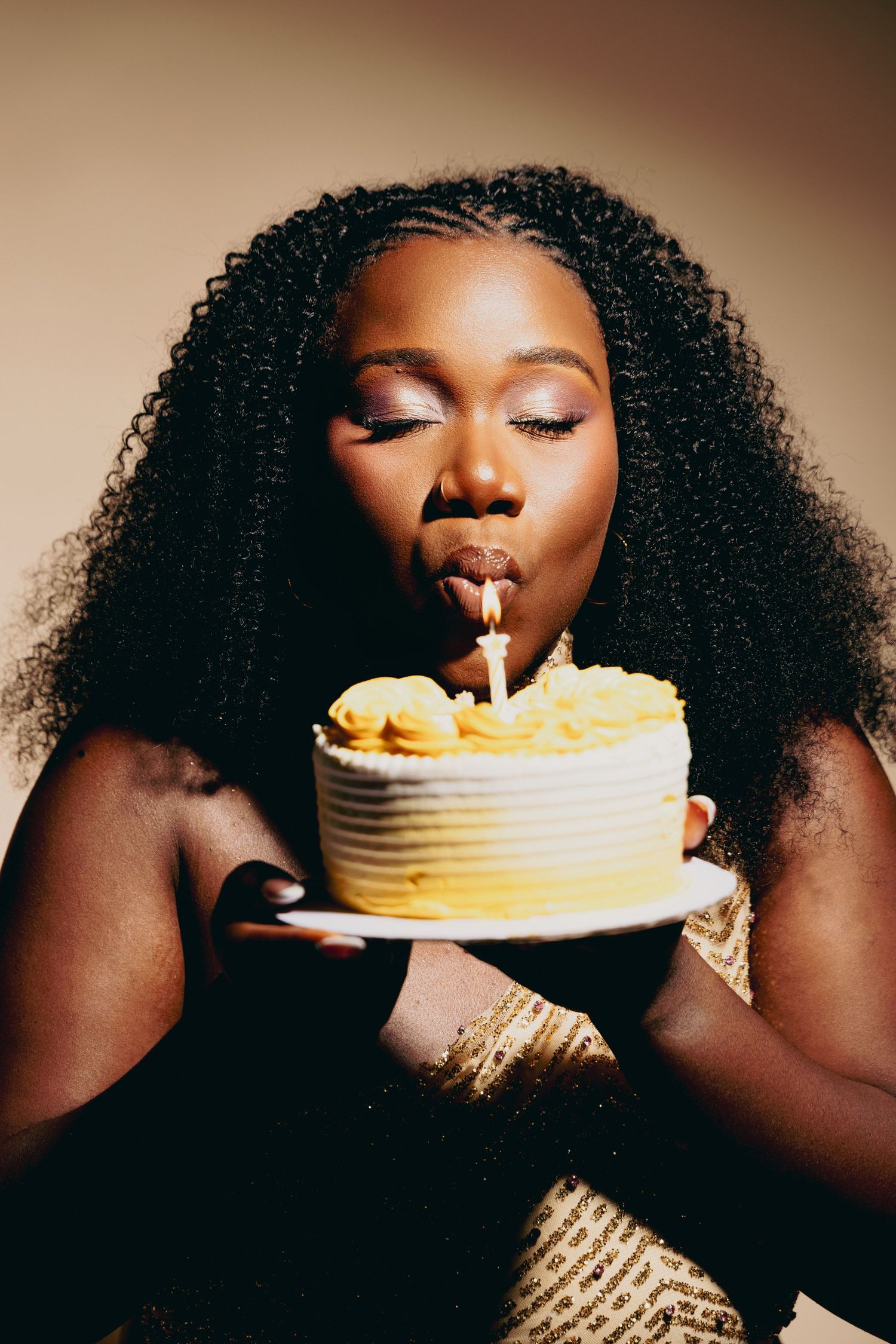 A young Black woman with curly hair blows out a birthday candle on a yellow ombre cake she holds on a plate.