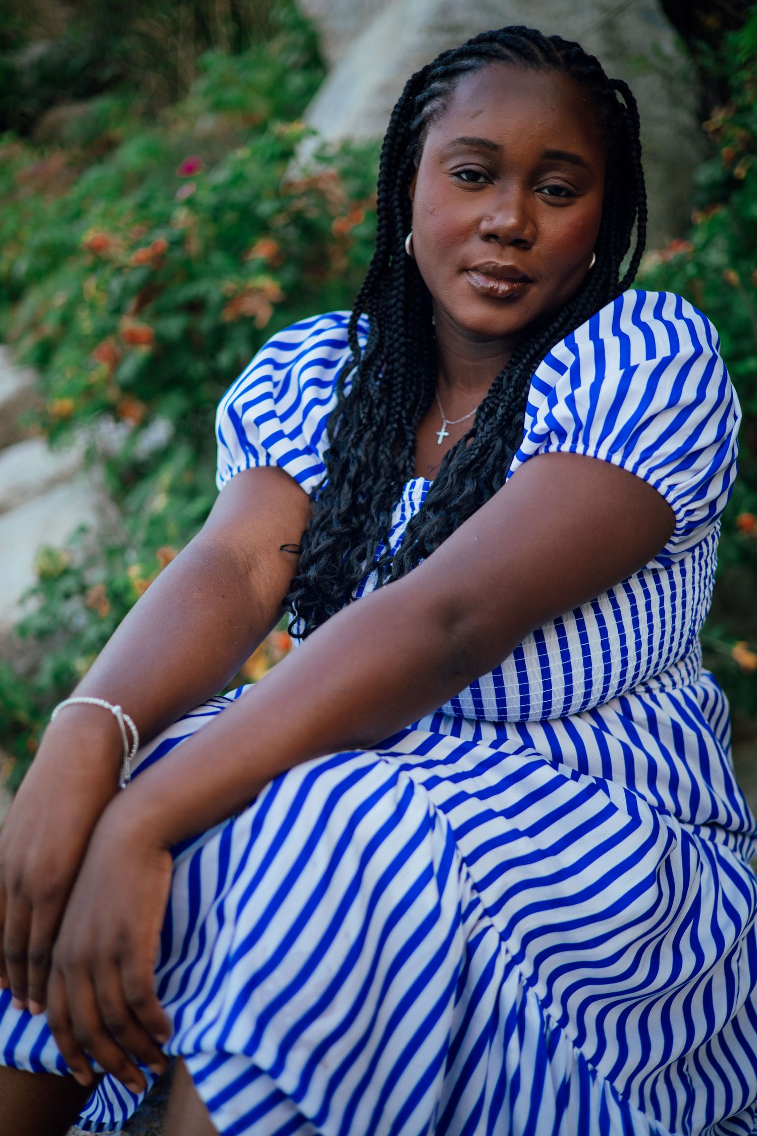 A young Black woman in a blue and white striped dress sits outdoors with braided hair, wearing a cross necklace.