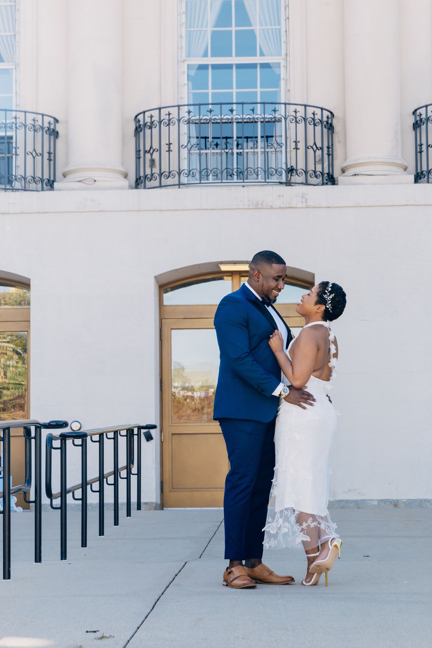 A bride and groom share a romantic embrace outside a neoclassical building with iron balconies.