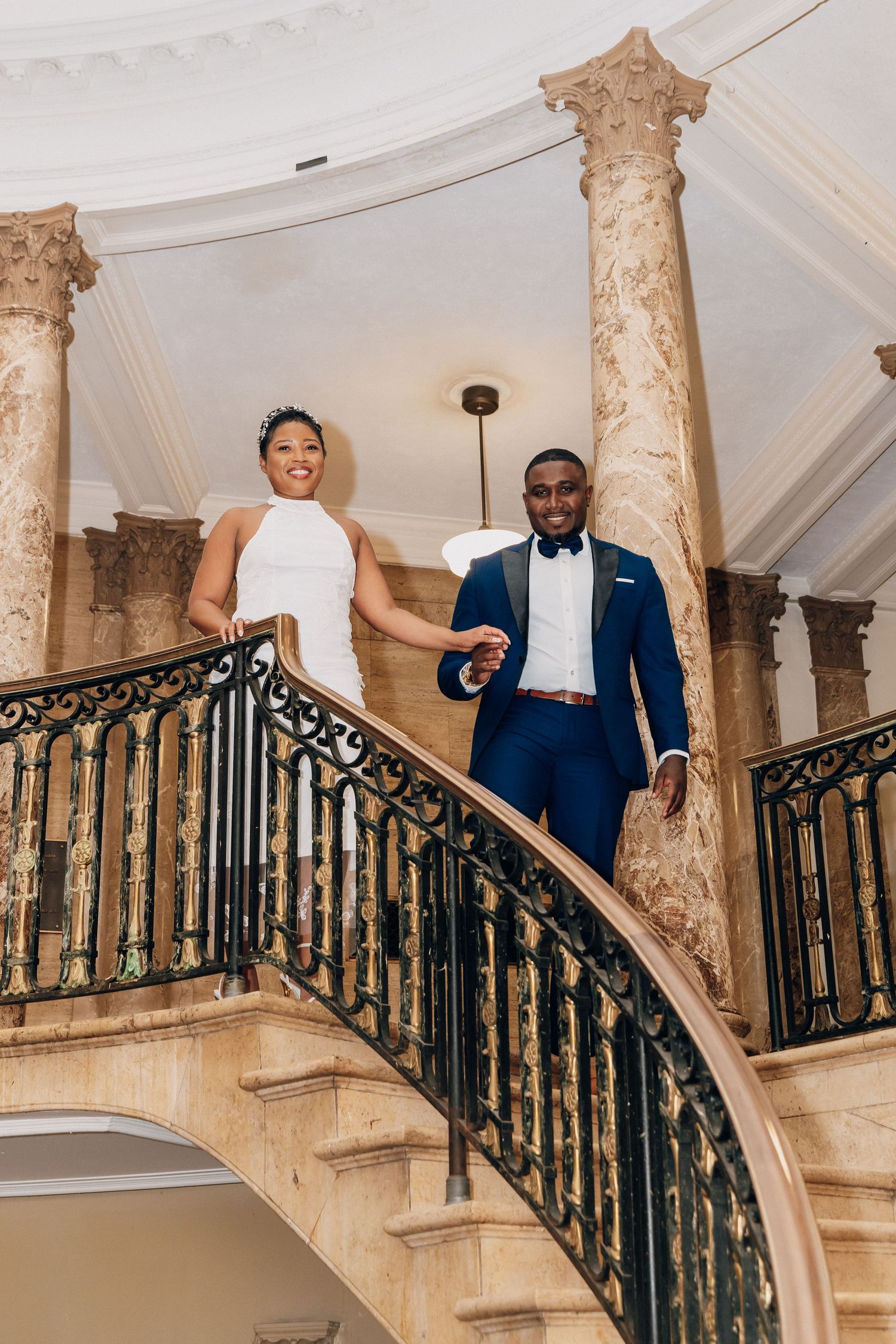 Bride and groom holding hands on an elegant curved staircase with ornate iron railings and marble columns.