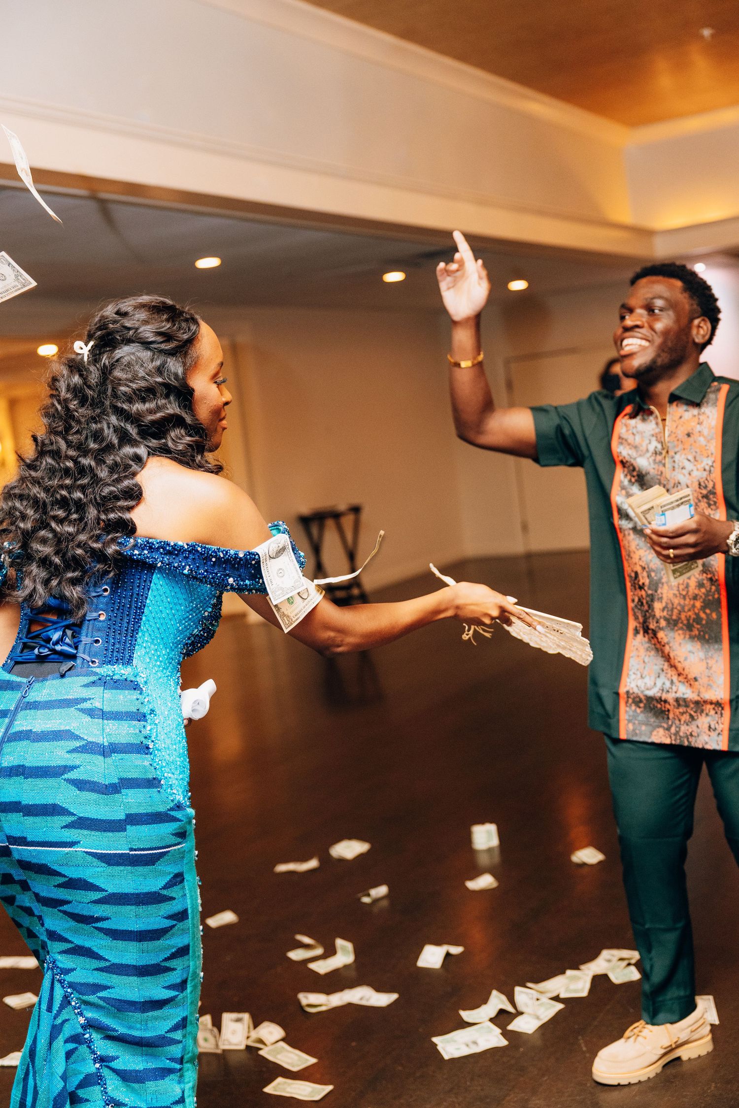 A joyful couple in African traditional wear celebrates by tossing money bills on a dance floor at a festive event.