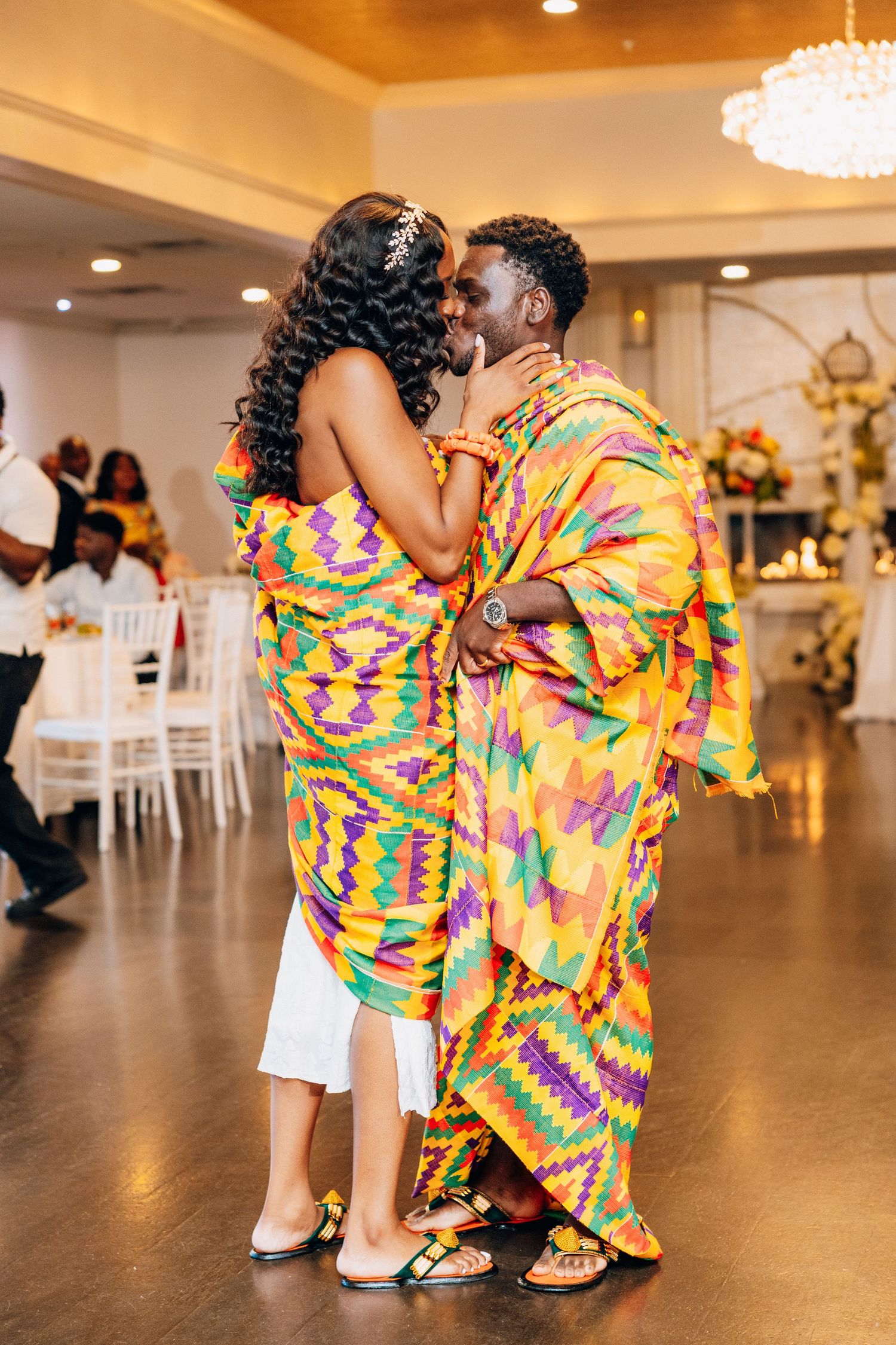 Couple sharing a kiss while dancing in matching colorful Kente cloth outfits at a wedding reception.