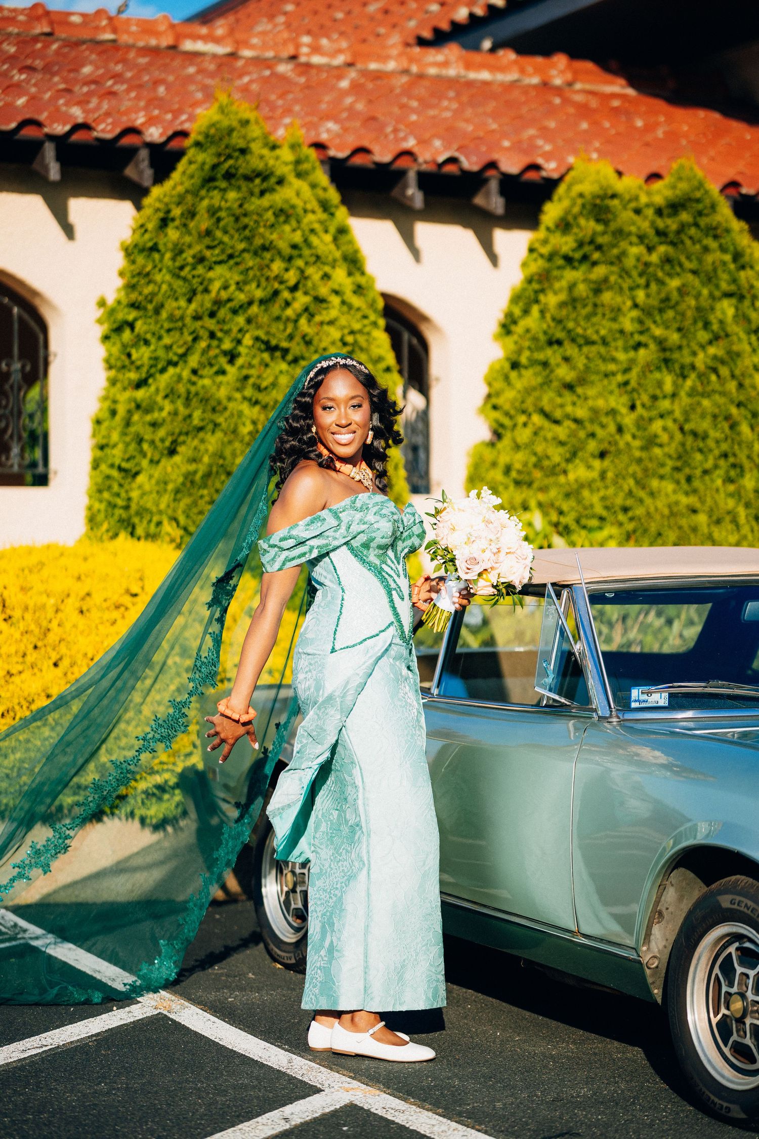 A Black bride in a teal off-shoulder gown with flowing veil holds a bouquet beside a vintage car.