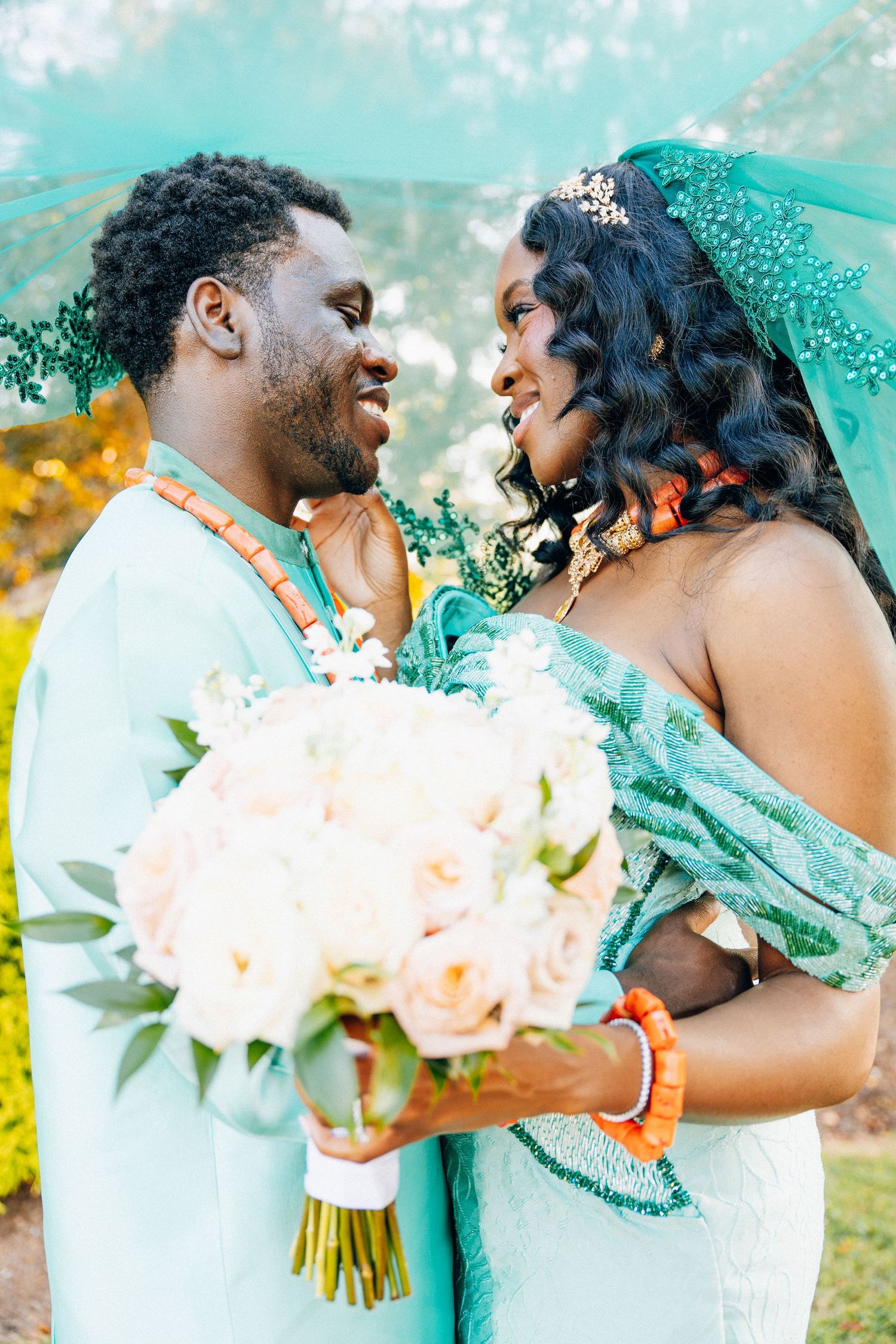 African couple in teal attire with orange beads share a loving gaze under veil.
