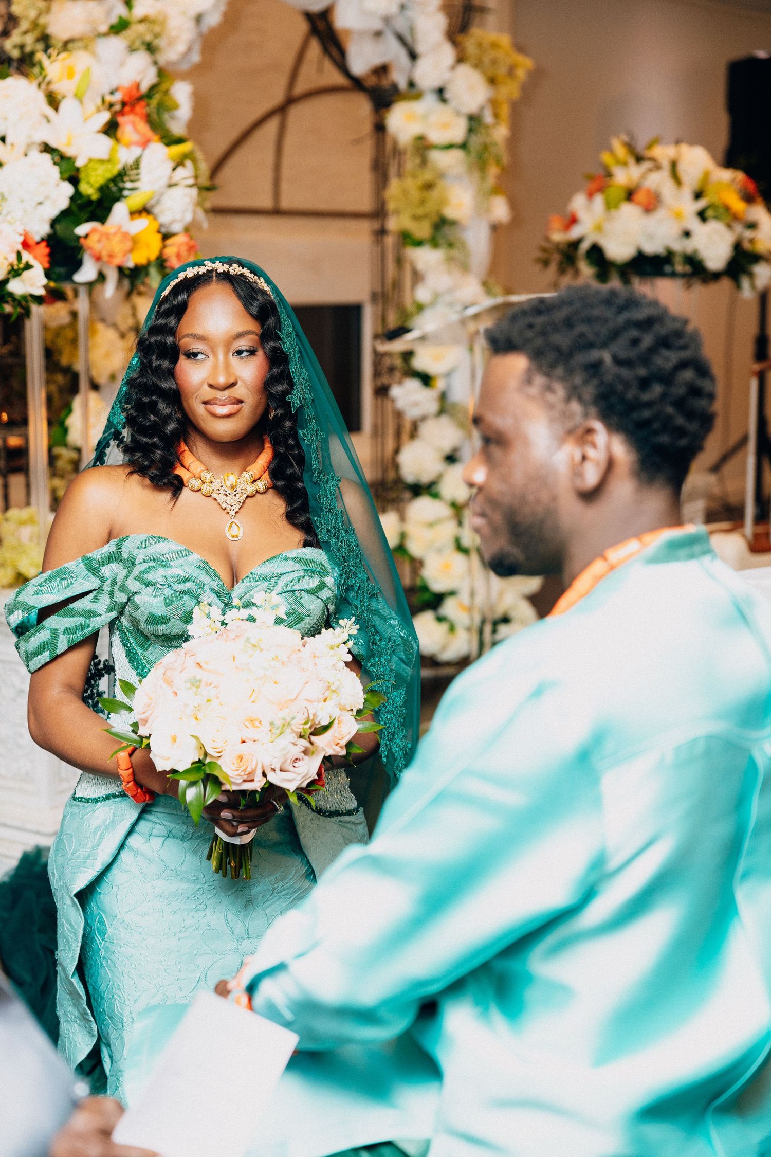 Bride in teal African gown with orange jewelry holds bouquet while gazing at groom during traditional wedding ceremony.