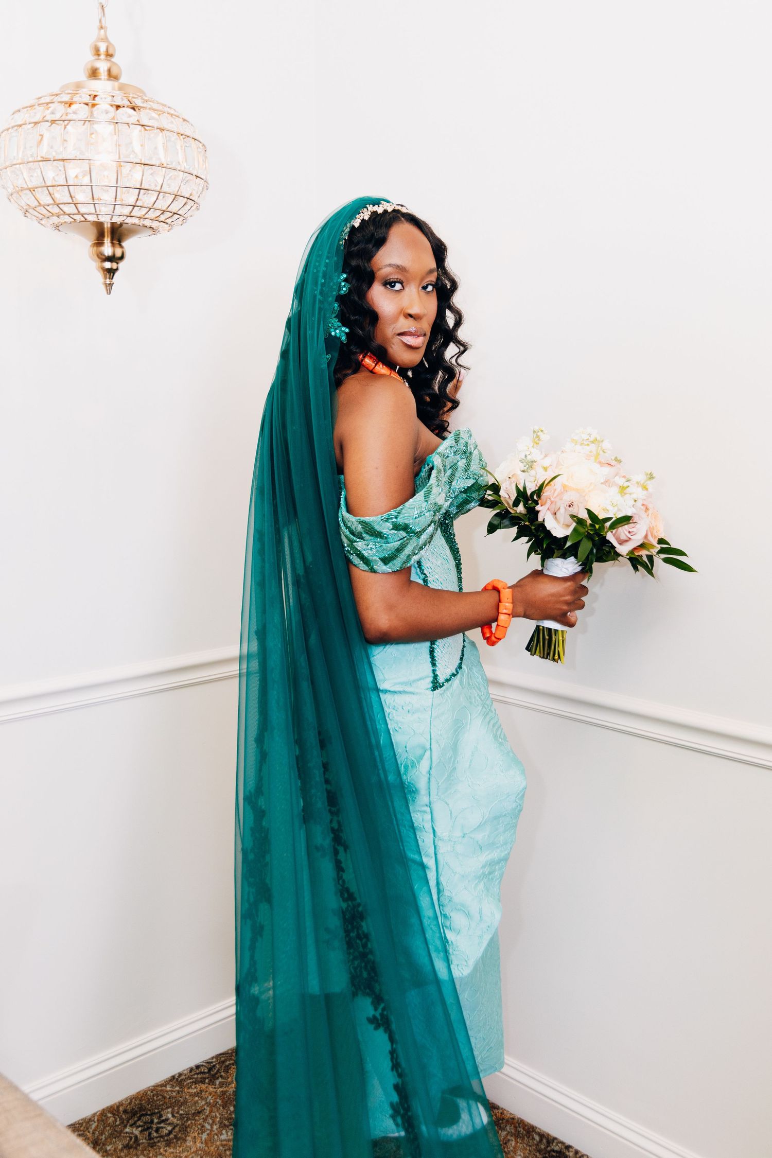 Bride in teal mermaid wedding dress with matching veil holding a blush bouquet under a gold chandelier.