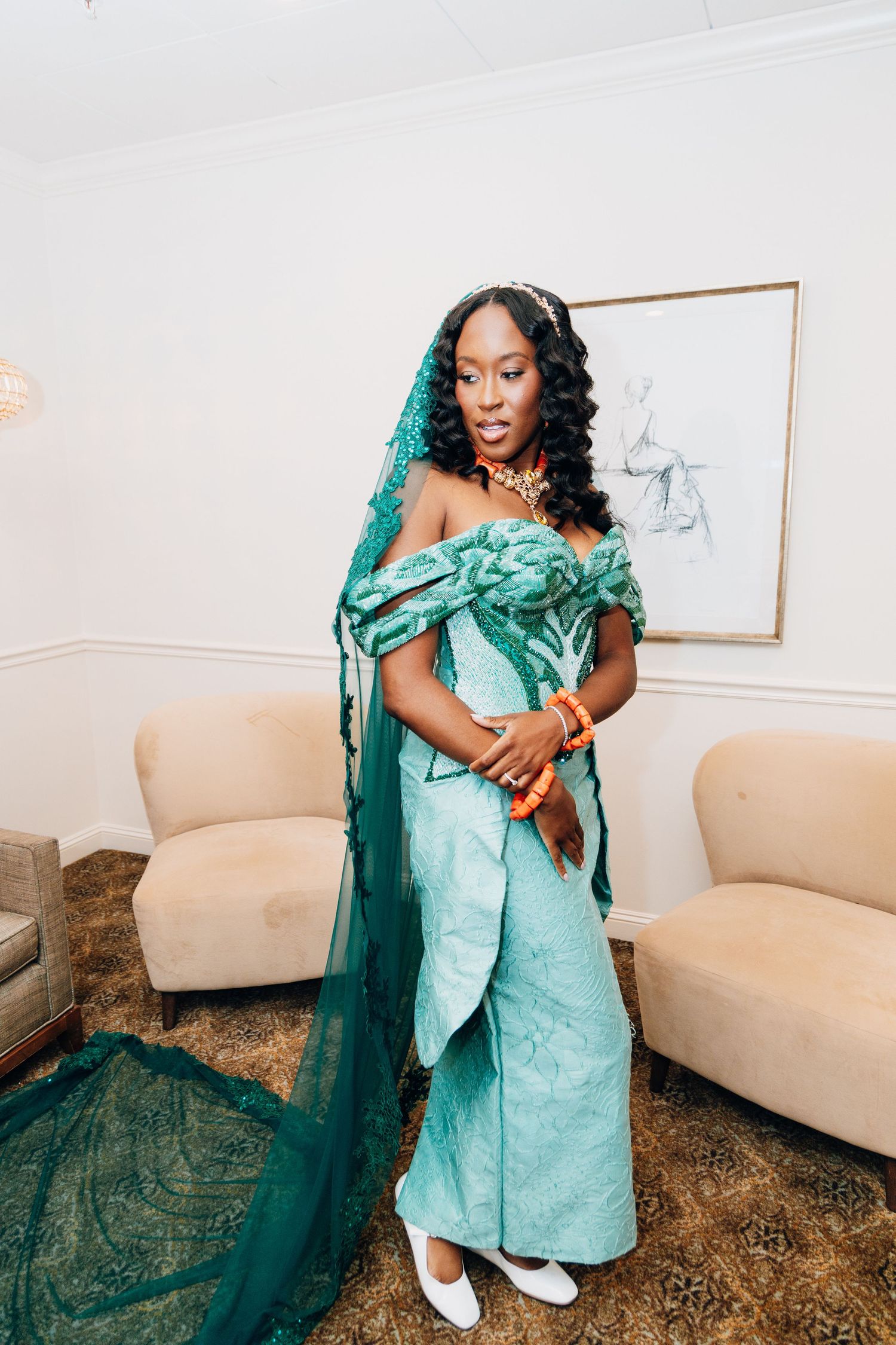 Woman in teal African bridal outfit with lace veil, orange jewelry, and white heels posing indoors.