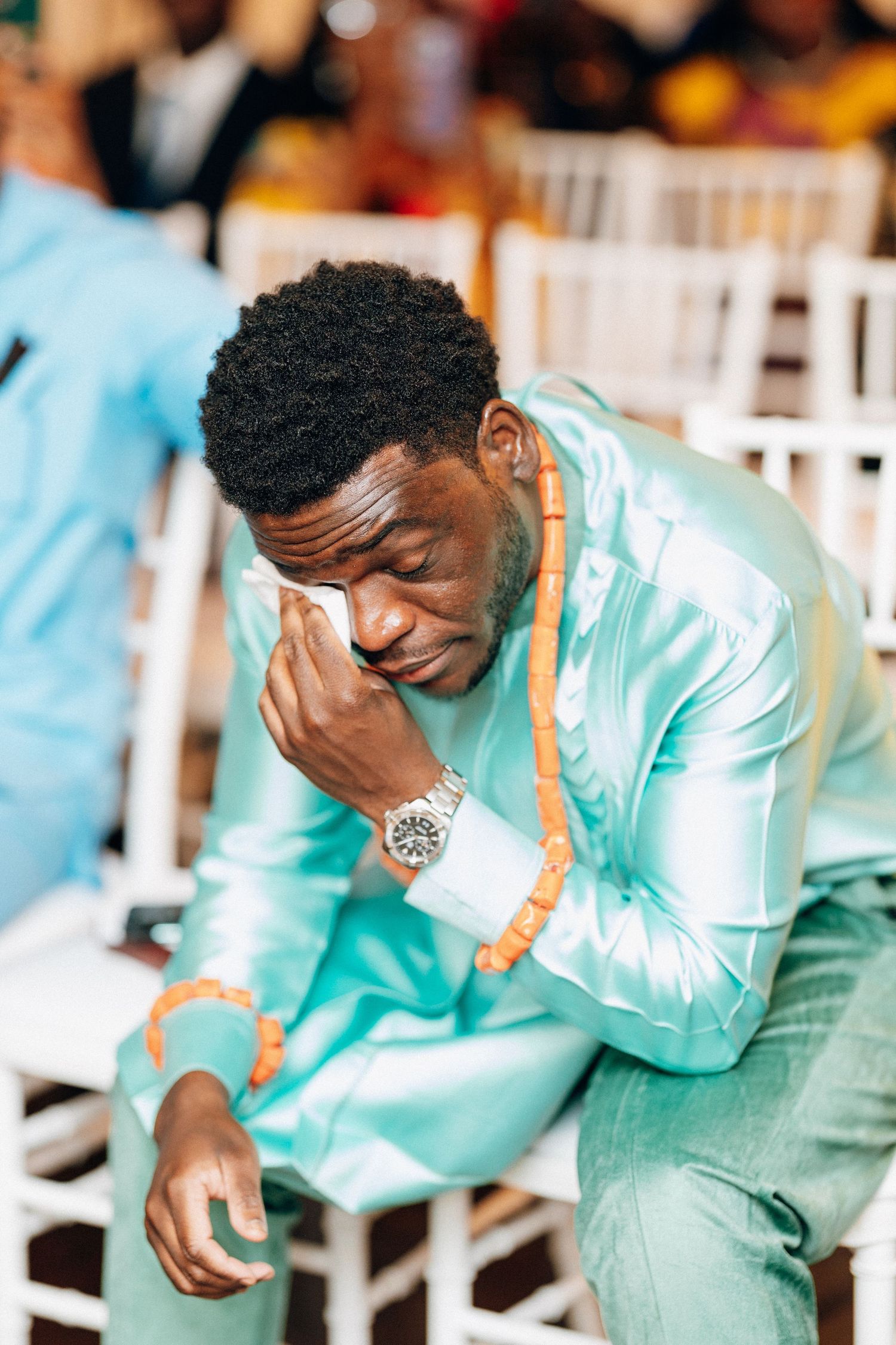 Emotional groom in teal satin traditional African suit wipes tears during wedding ceremony.