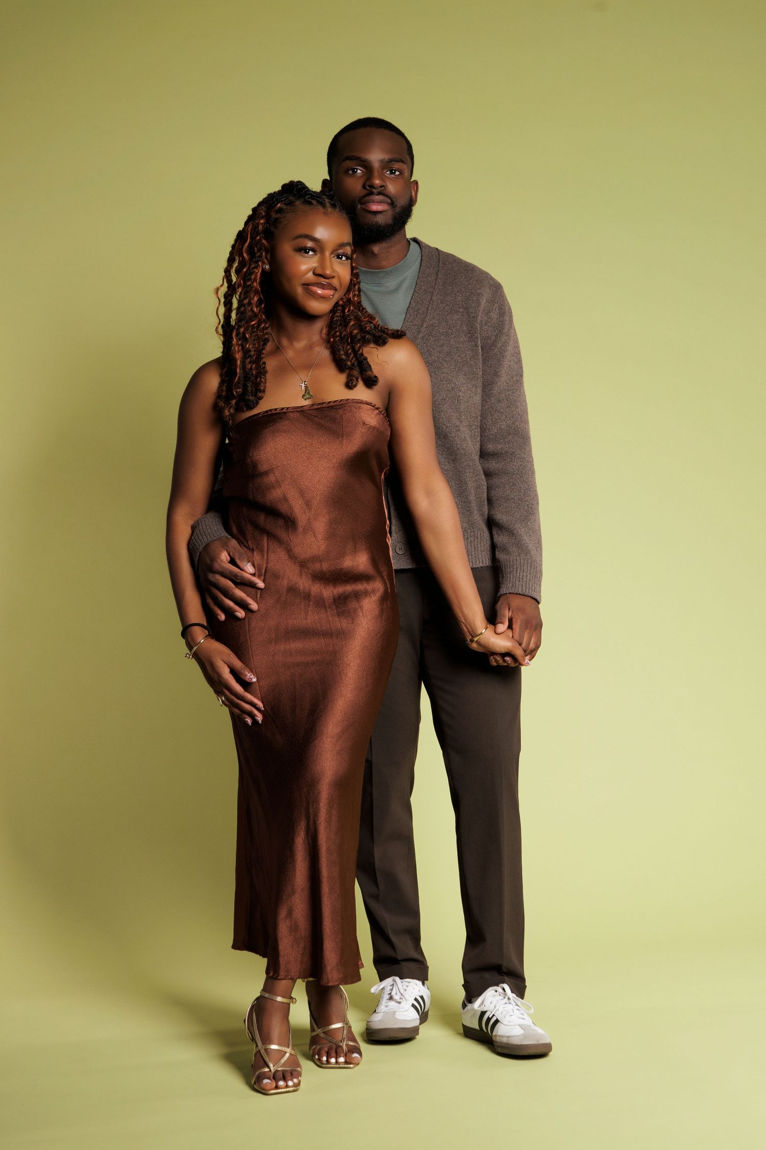 A pregnant Black woman in a brown satin slip dress poses with a man in a gray cardigan against a green background.