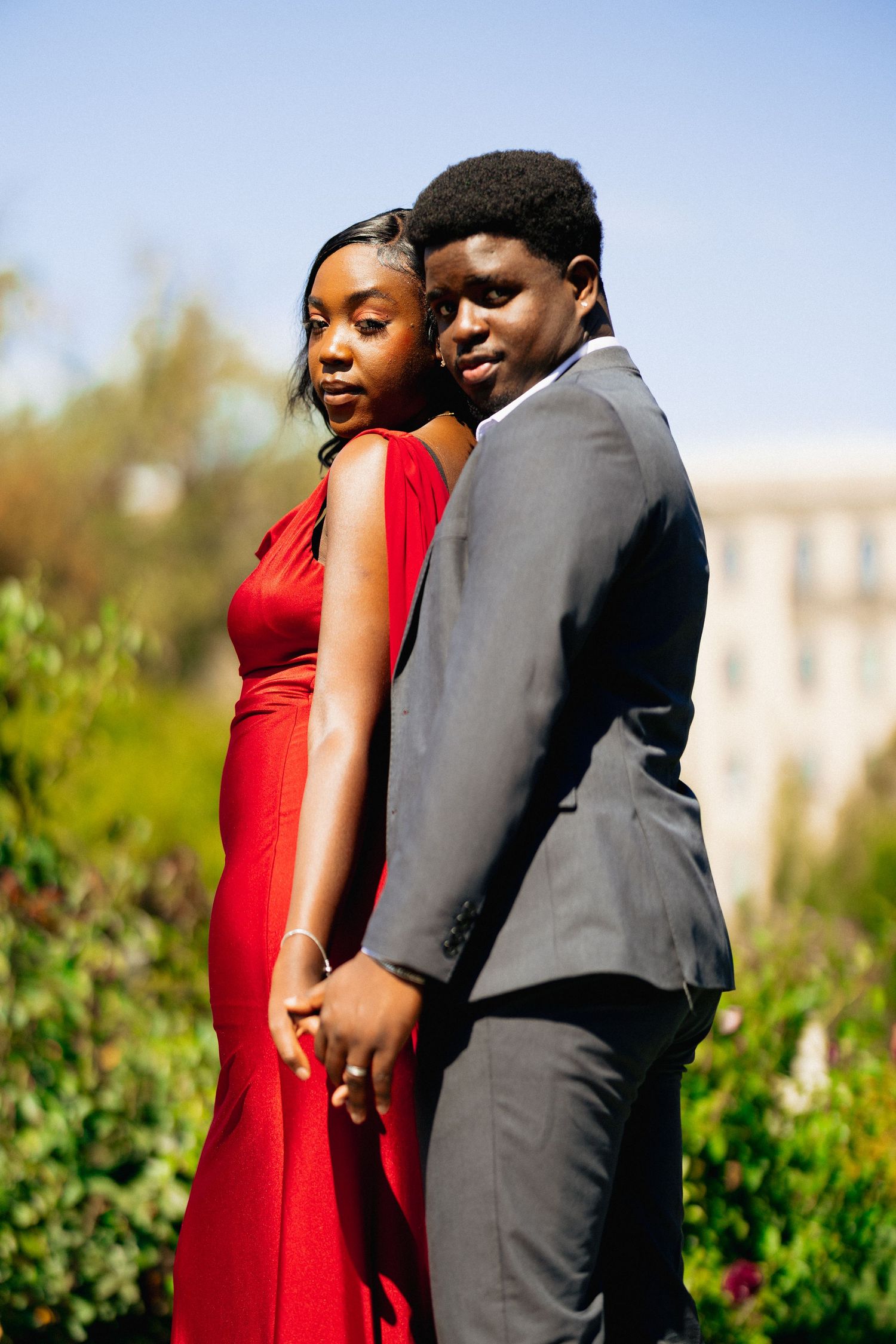A couple poses back-to-back outdoors, woman in red dress and man in gray suit looking over shoulders.