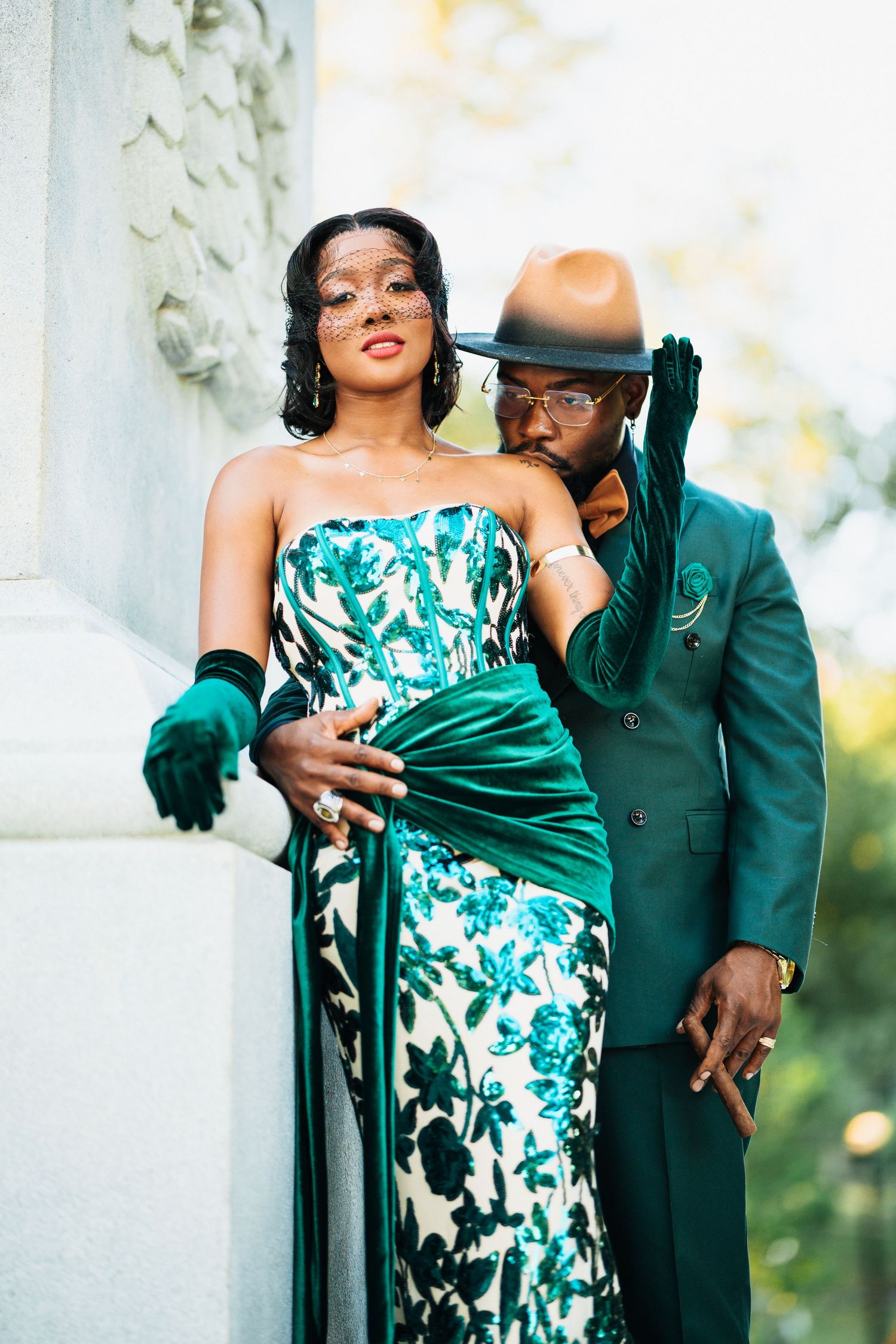 Glamorous couple in green floral gown and teal suit posing by ornate stone column.