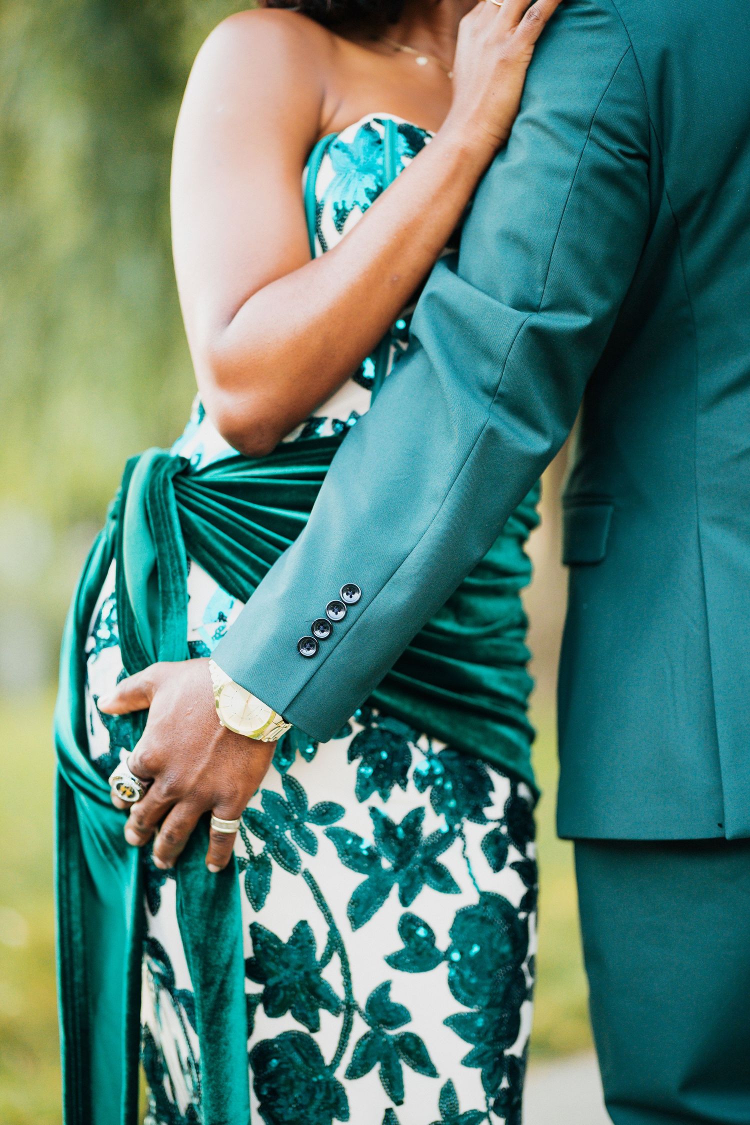 Close-up of Black couple embracing, showcasing green floral gown and teal suit with gold watch and rings.