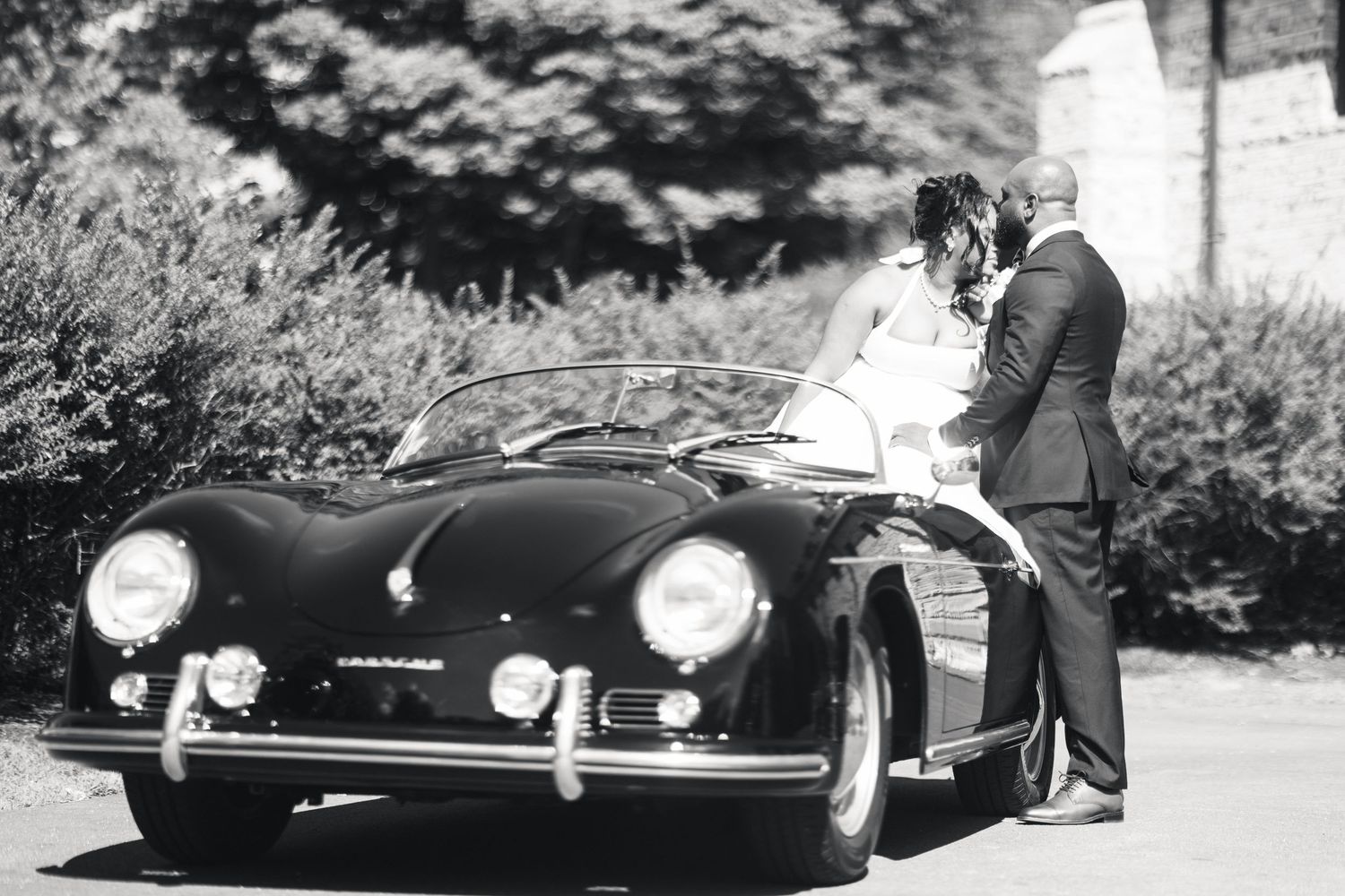 Black and white photo of couple kissing beside a classic vintage Porsche speedster outdoors.