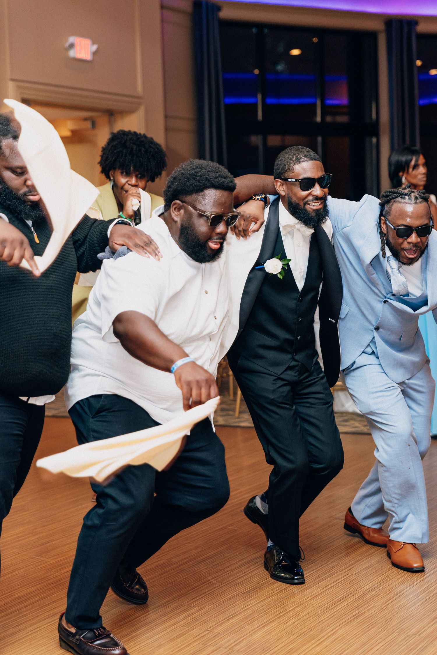 Groom and groomsmen dancing energetically at wedding reception, wearing suits and sunglasses in festive celebration.