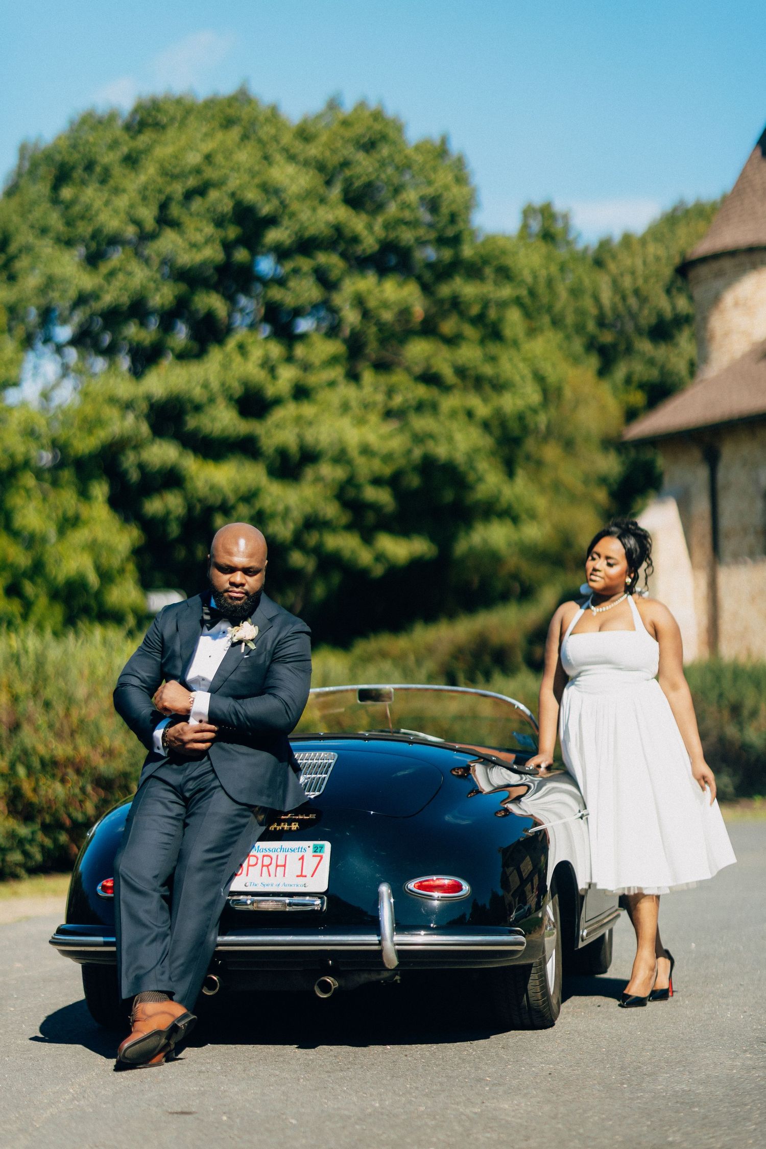 Elegant bride and groom posing beside a vintage black Porsche 356 convertible on wedding day.