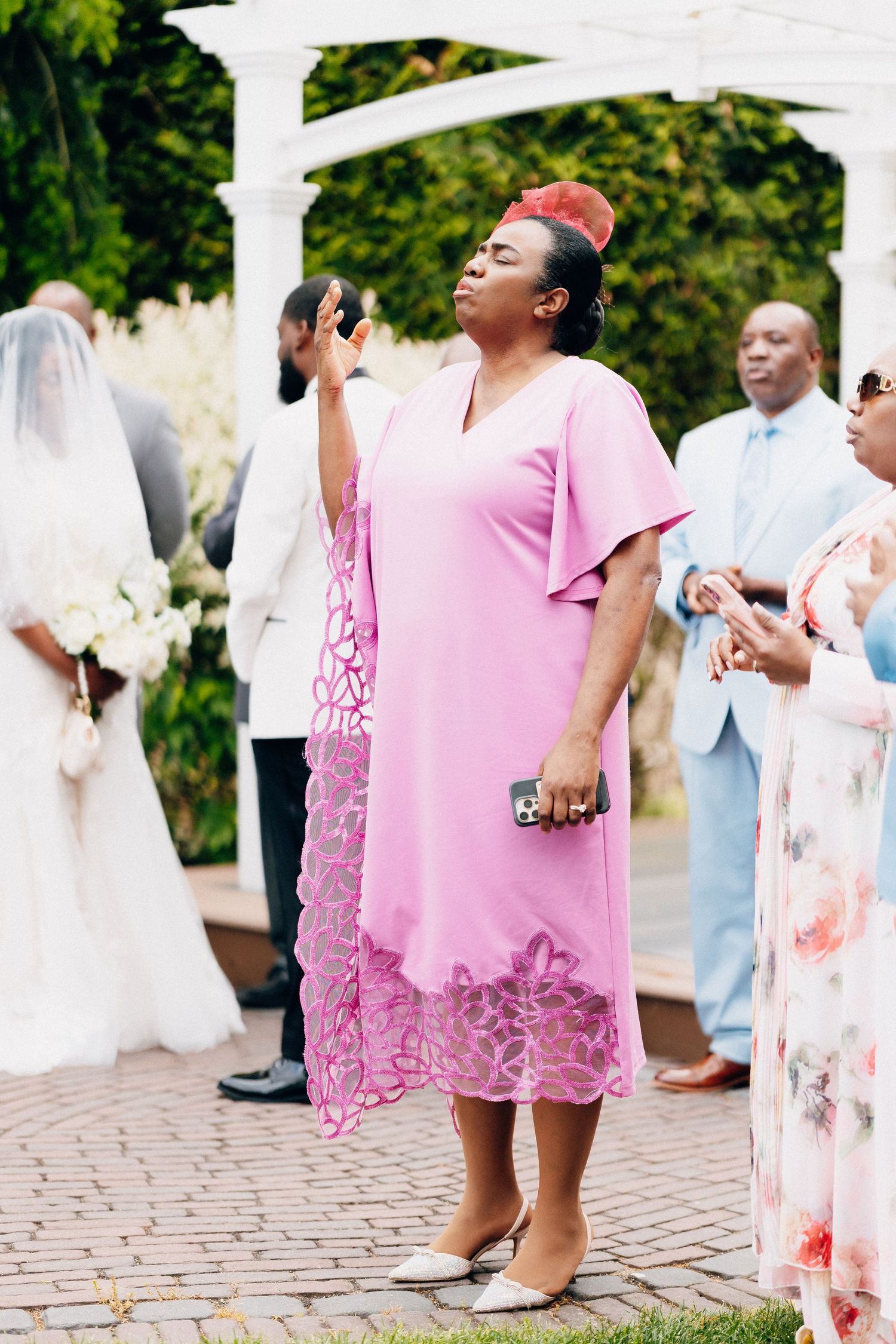 Woman in pink dress raises hand emotionally during outdoor wedding ceremony near white pergola.