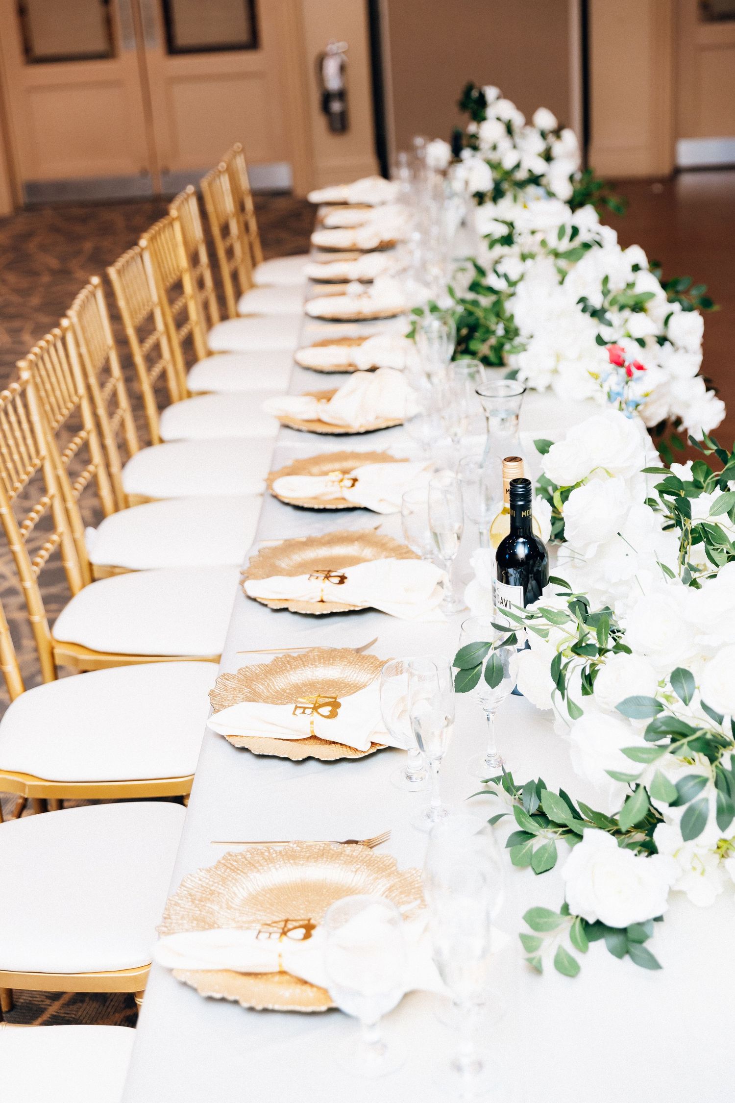 Elegant wedding reception table with gold charger plates, white floral centerpieces, greenery runner, and gold chiavari chairs.