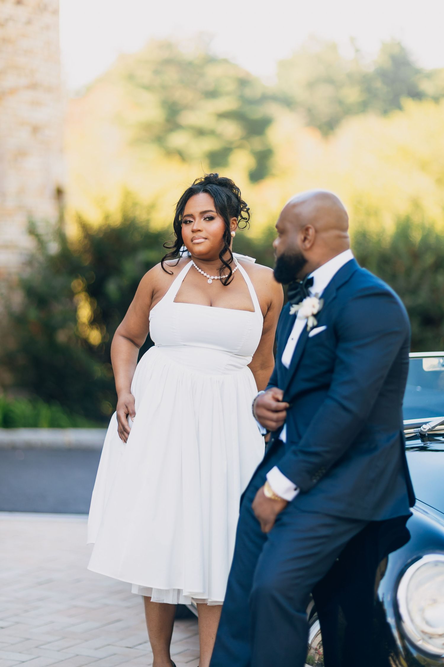 A bride in a white halter dress and groom in navy suit pose by a classic black car outdoors.